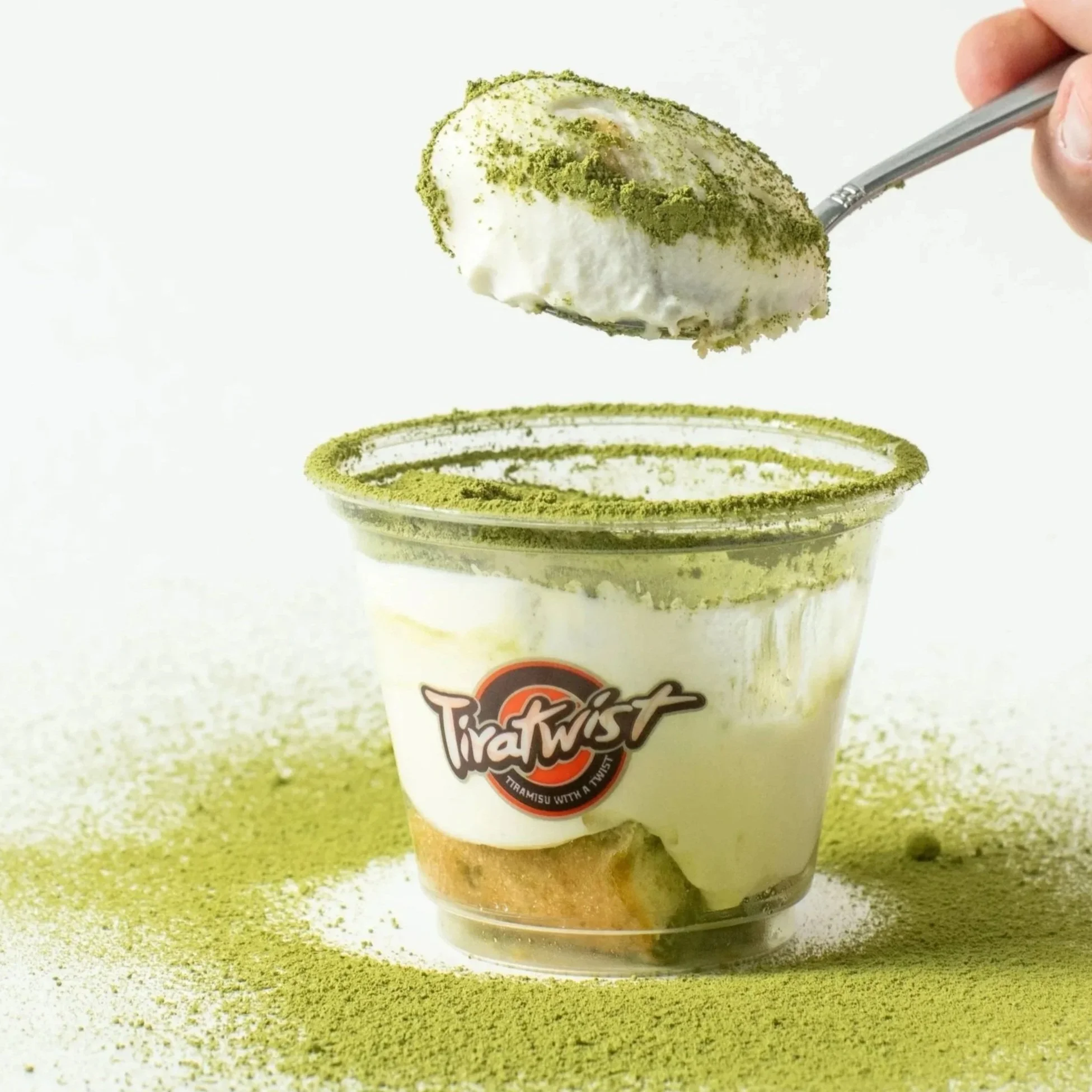 A cup of tiramisu with a creamy topping dusted with green matcha powder, with a hand holding a spoon above it, also coated with matcha powder.
