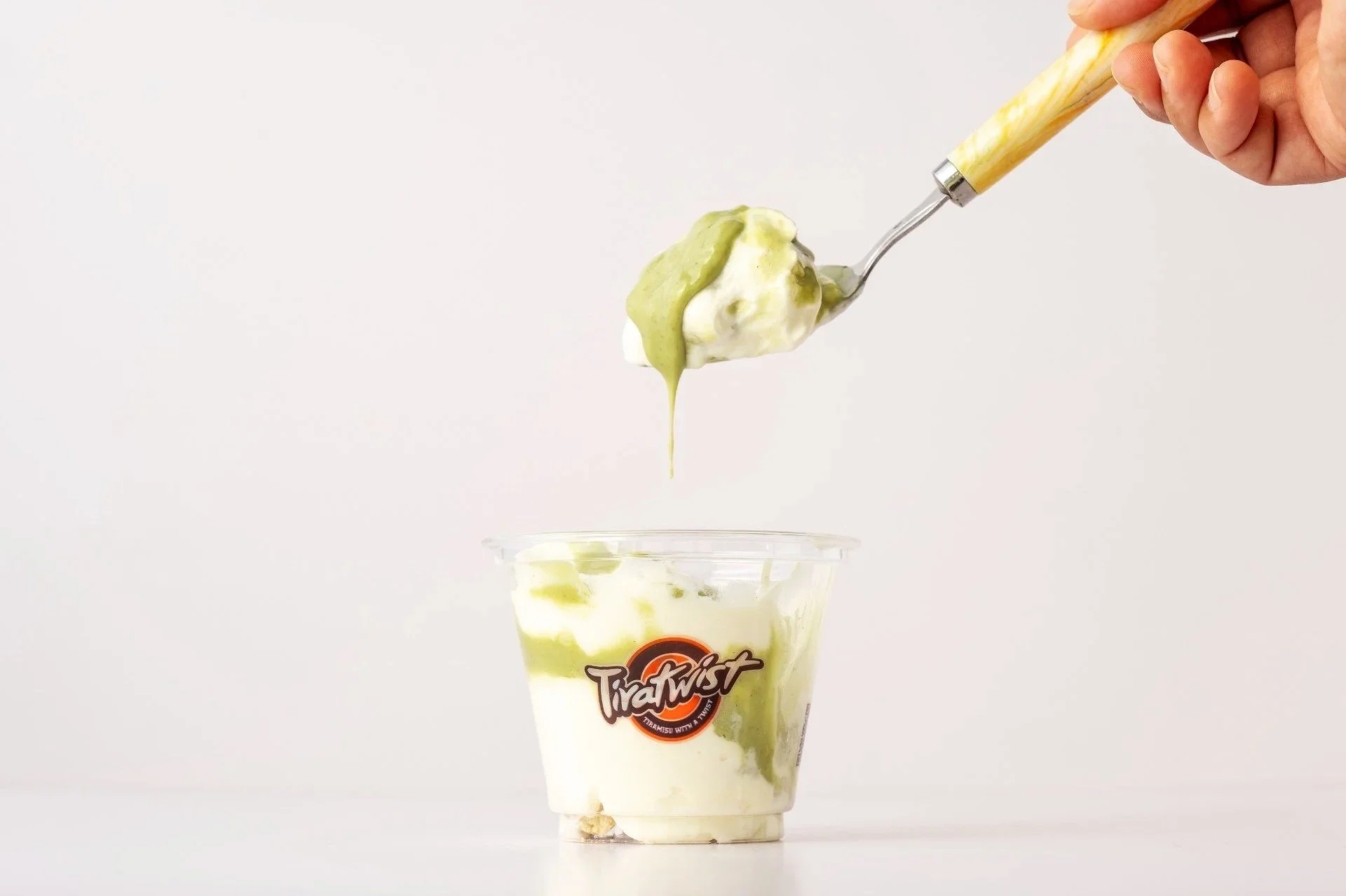 A person is scooping pistachio tiramisu with a yellow marble-patterned spoon into a clear plastic cup labeled