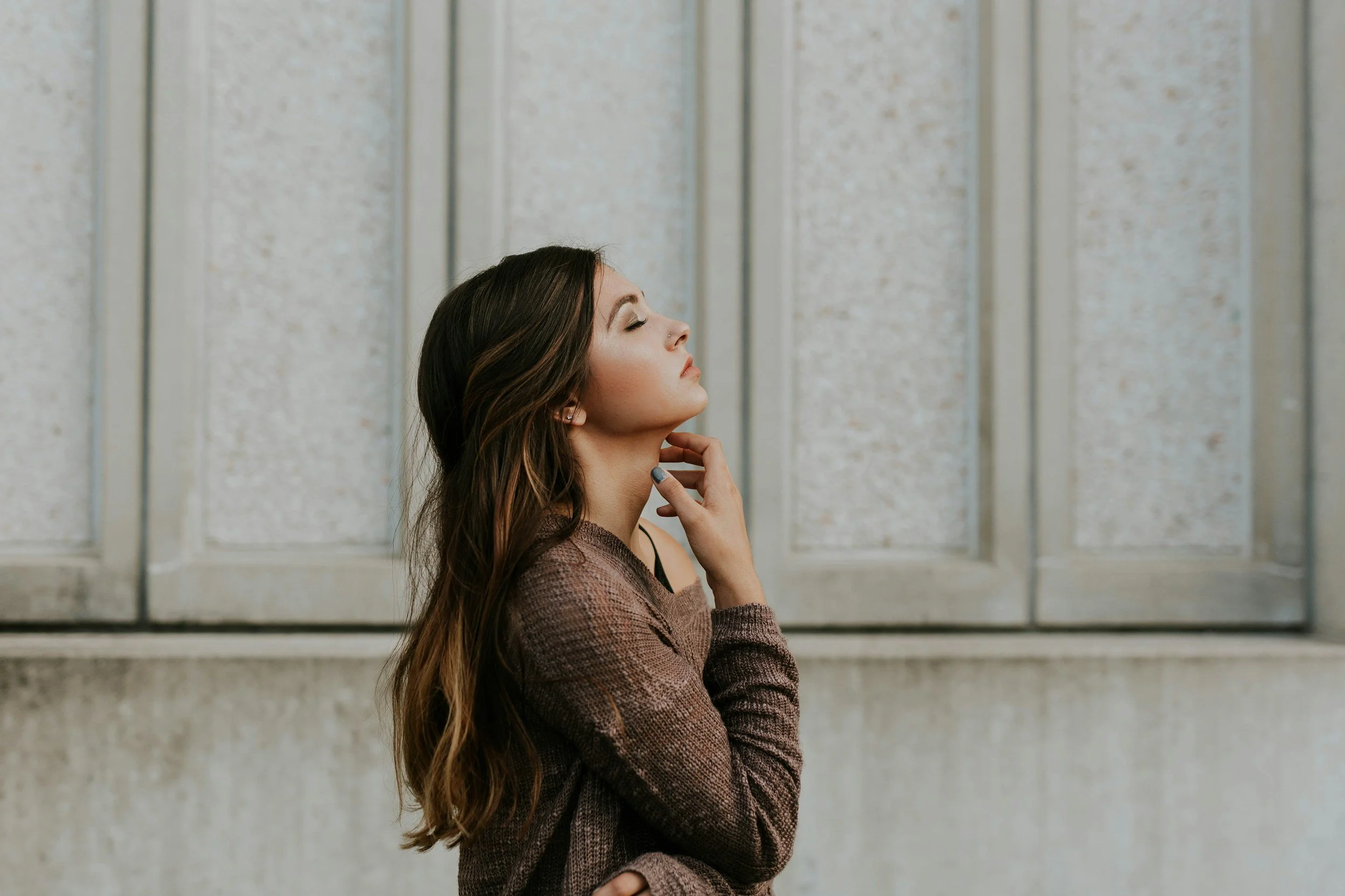 Side profile of a woman with long brown hair, eyes closed, standing against a textured gray wall.