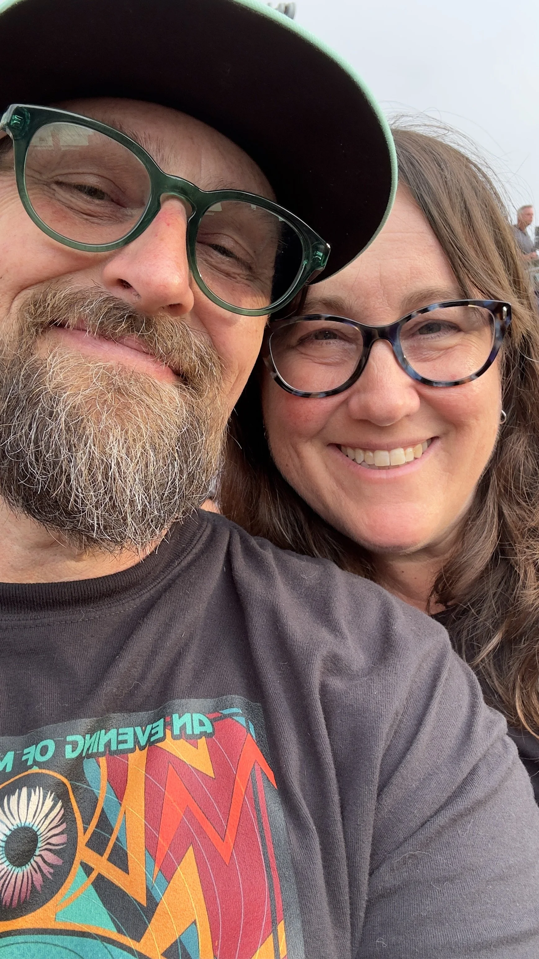 A close-up of a smiling couple taking a selfie. The man has a beard, wears glasses and a baseball cap. The woman has long hair, glasses, and is smiling. There are people in the background at an outdoor day event.