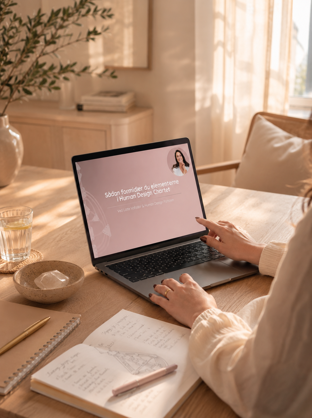 Person sidder ved et træbord med en åben MacBook, der viser et præsentationsslide om Human Design Chart. Der er en notesbog, en pen, en kop vand, en skål med en klump, og et åbent sketchbook med tegninger. Sollys skinner gennem vinduet.
