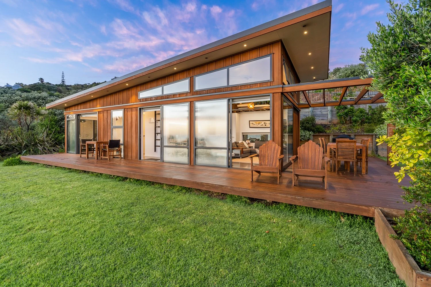 Modern house with large glass doors and a wooden deck, outdoor seating, green lawn, and a hillside with trees in the background during sunset.