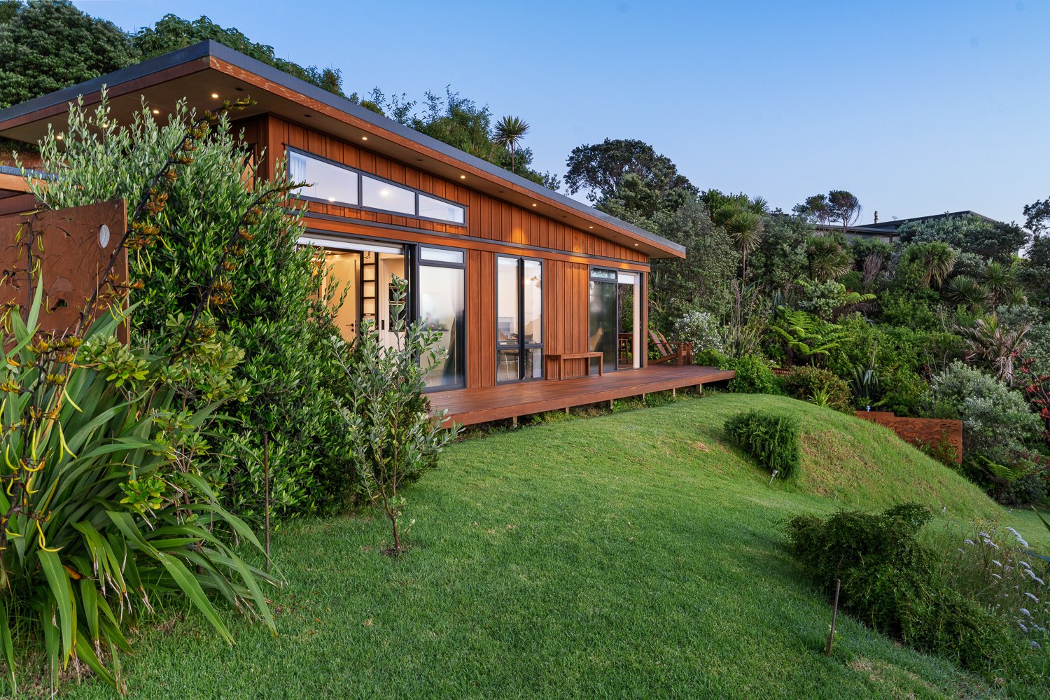 Modern wooden house with large glass windows and sliding doors, situated on a green hillside with lush plants and trees surrounding it.