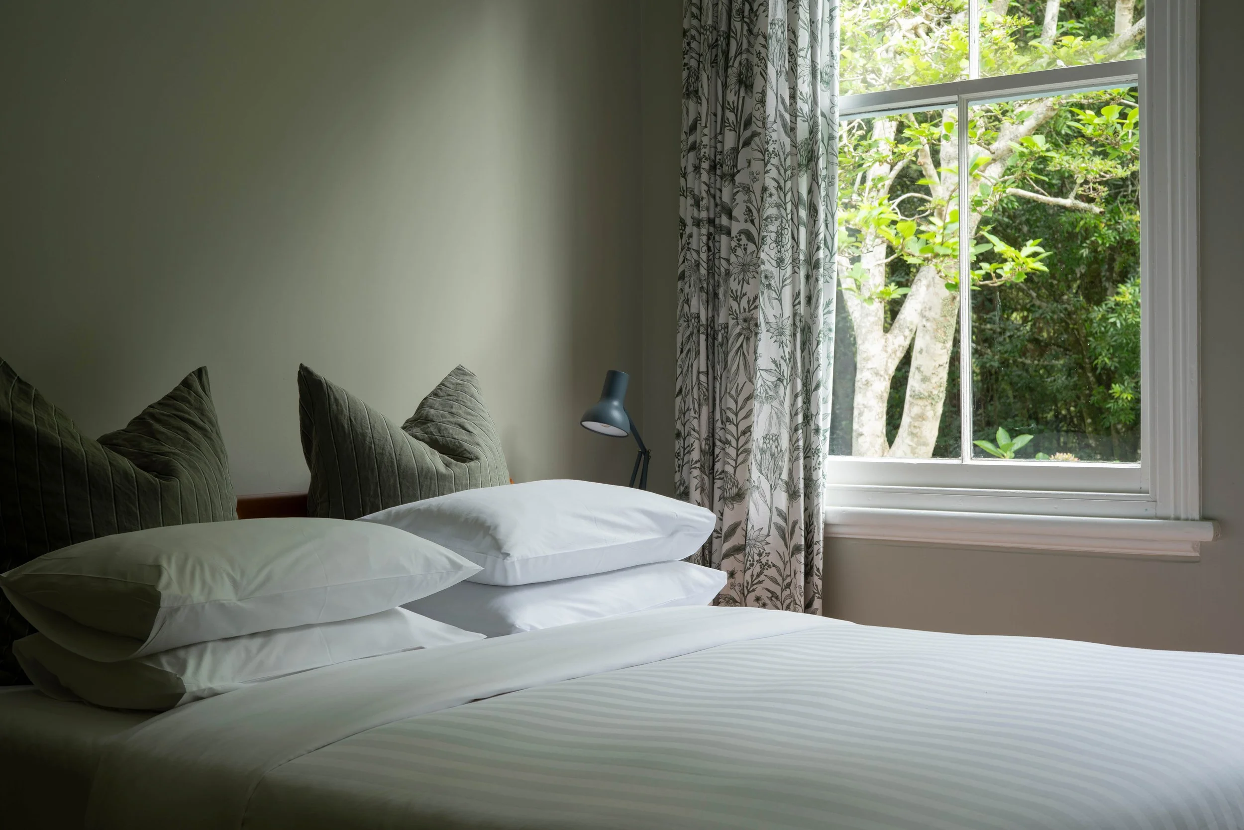 A bedroom with a bed, white bedding, four pillows, two dark green pillows, and a bedside lamp next to a window with floral curtains, overlooking trees outside.