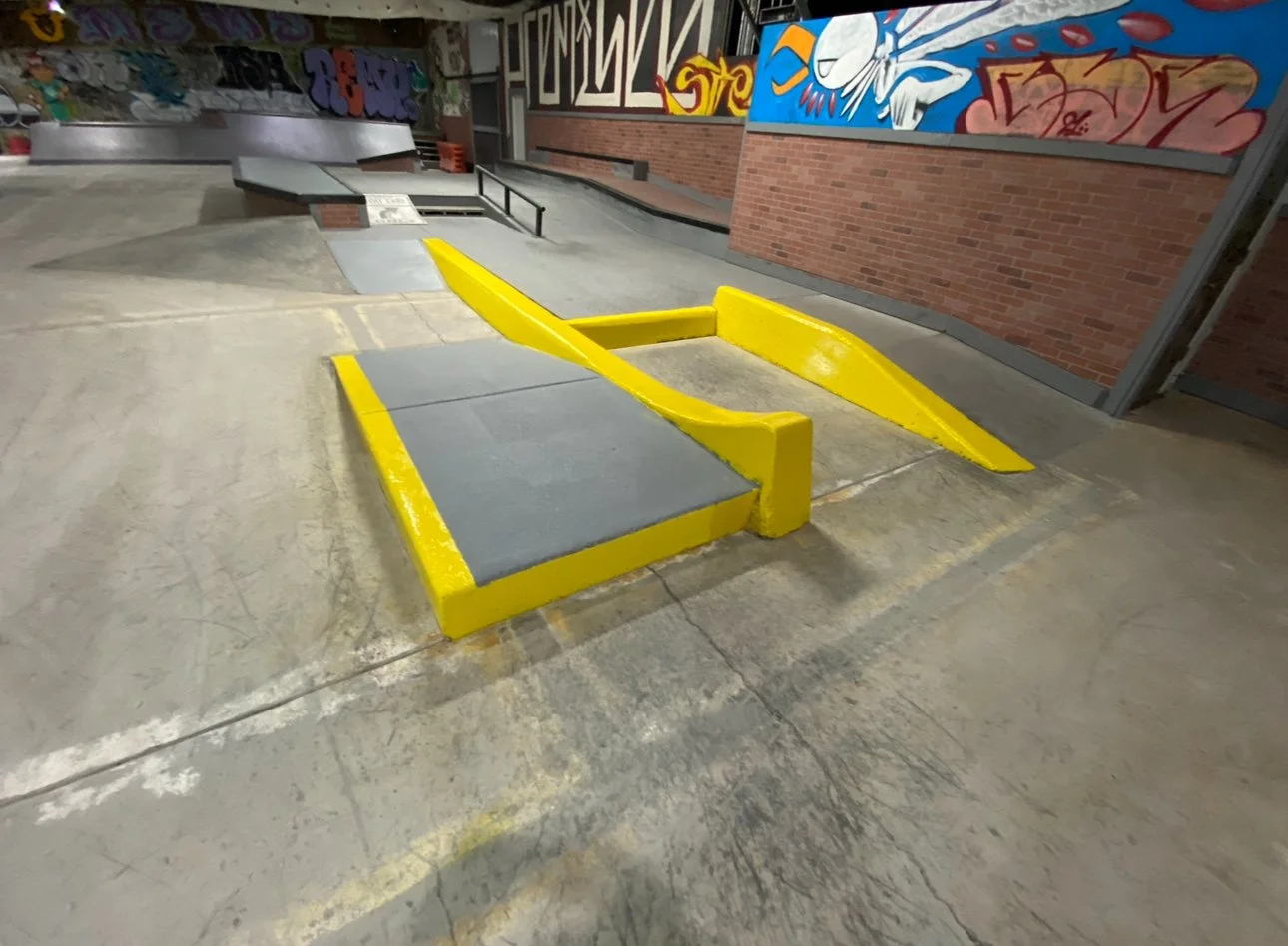 Empty indoor skatepark with yellow-painted ramp and ledge, graffiti art on brick wall.