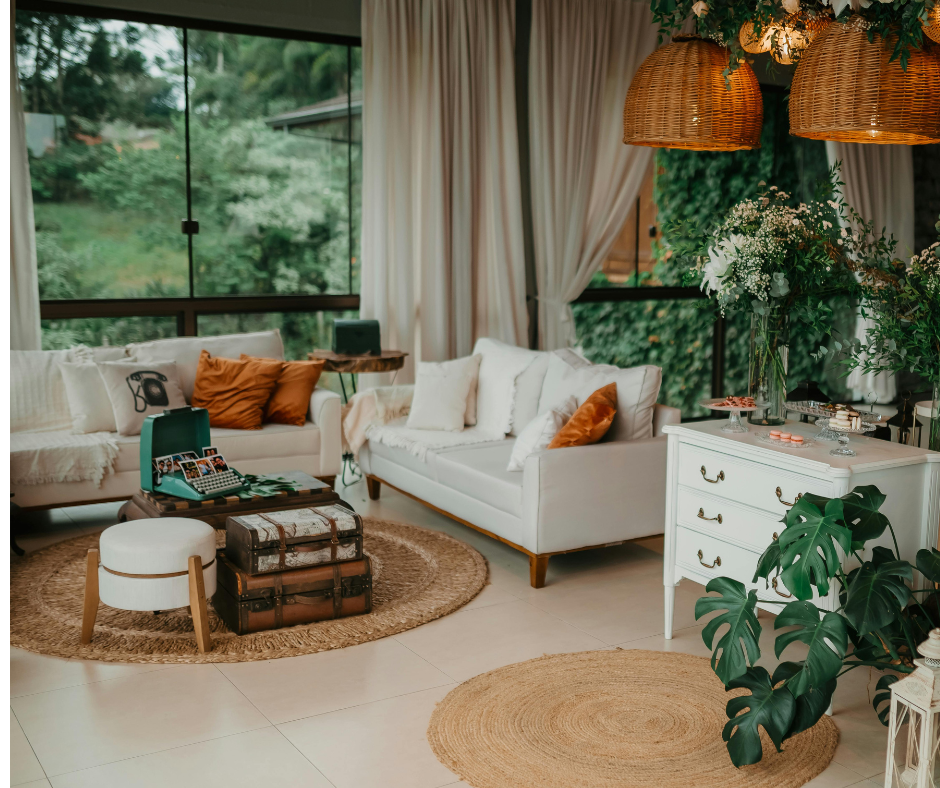 A cozy living room with white couches, decorative pillows, a circular woven rug, large windows with curtains, and greenery outside. Decorative items include a vintage suitcase, a small white ottoman, a white dresser with flowers on top, and hanging wicker lamps.