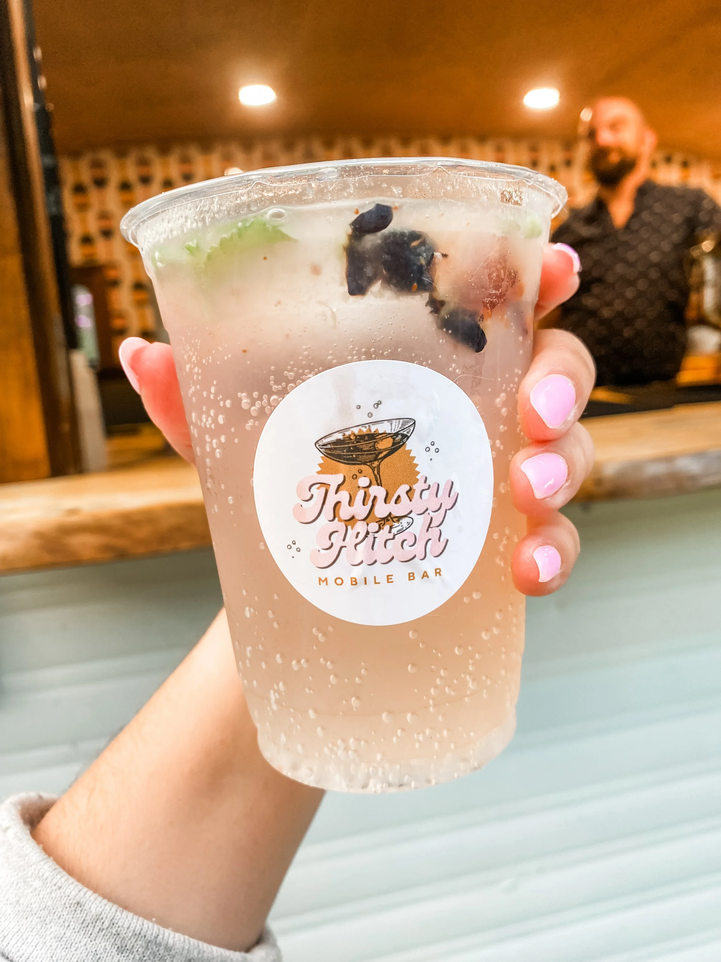 A woman holding a cocktail with a bubbly drink, garnished with blueberries and mint, in front of a vintage bar trailer. The Thirsty Hitch mobile bar and bartending serves Haliburton, Kawartha Lakes, Muskoka, and beyond!
