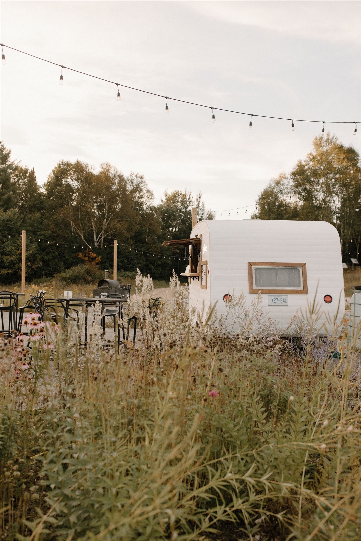 A small white vintage bar trailer set in a field of wildflowers with string lights hanging above, surrounded by trees.  The Thirsty Hitch mobile bar and bartending serves Haliburton, Kawartha Lakes, Muskoka, and beyond!