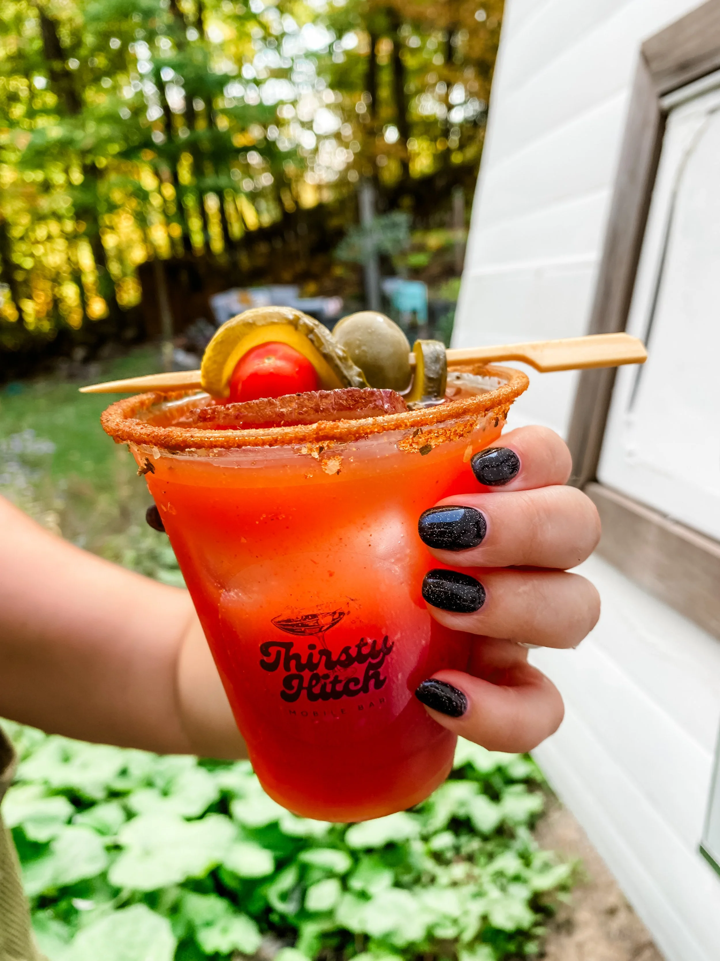 A hand holding a red cocktail in a plastic cup with the logo 'Thirsty Hitch Mobile Bar'. The cocktail has a salted rim and garnished with a large green olive, a cherry tomato, a pickle, and a lemon wedge on a skewer, set outdoors with trees and a white building in the background.