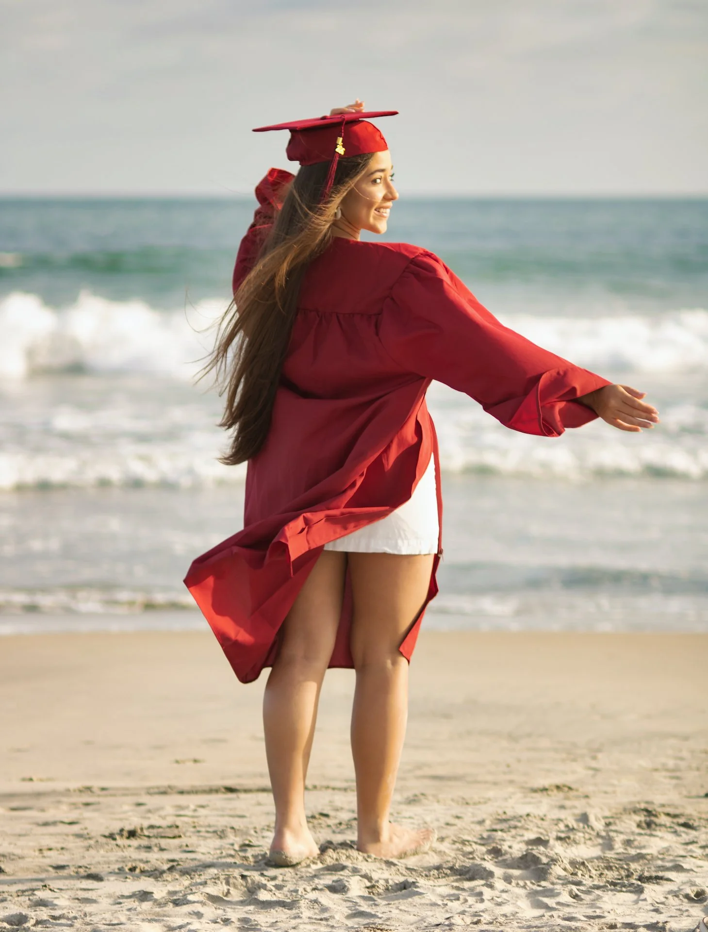 A young woman in a red graduation cap and gown standing on a sandy beach, smiling and looking over her shoulder at the ocean.