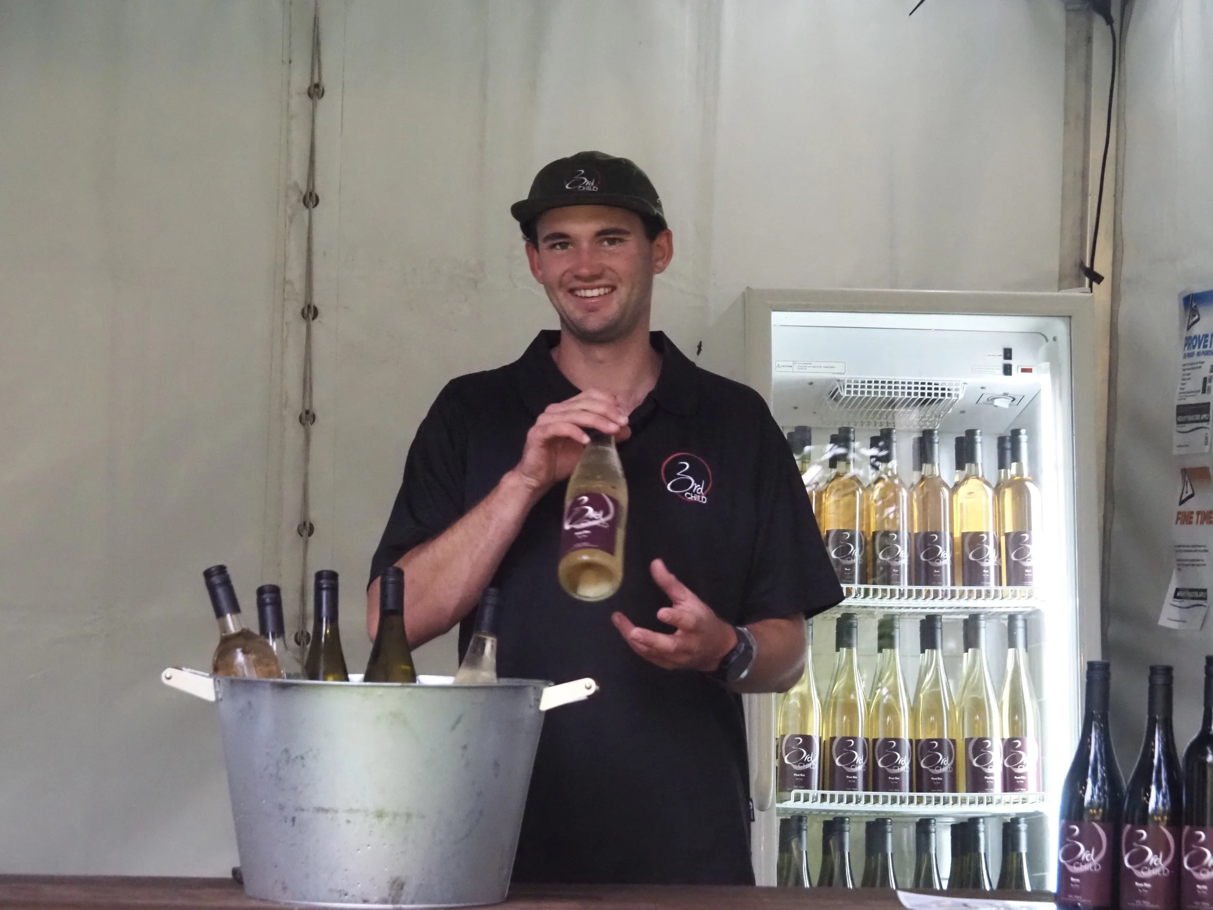 A young man in a black polo shirt with a logo and a black cap, standing behind a table with bottles of wine, holding a bottle and smiling at the camera. There is a refrigerator filled with more bottles of wine in the background. Coal river. Tasmania.