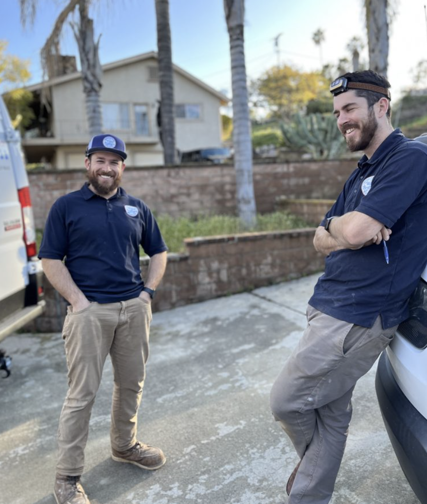 Two men wearing work uniforms, one with a cap and the other with a headlamp, stand and talk outside near a van and a house. Palm trees are in the background.