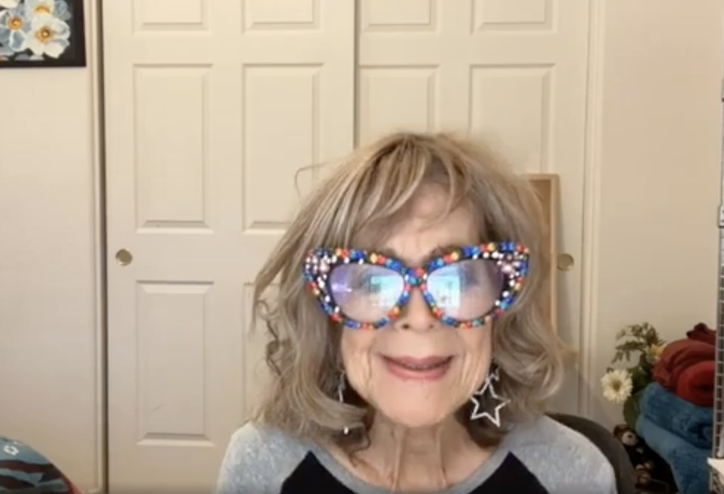 A woman with blonde hair and star-shaped earrings wearing colorful, sparkly sunglasses, smiling in a room with a white paneled closet door in the background and a floral arrangement on the right.