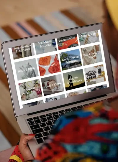 Person holding a laptop displaying an online shopping website with categories such as Accessories & Apparel, Art, Music & Books, Automotive, Baby & Kids, Eco-Friendly Goods, Bath & Body, Beauty, Commercial Services, Home Decor, Flowers, Party & Gifts, Food & Drink, Health & Wellness.