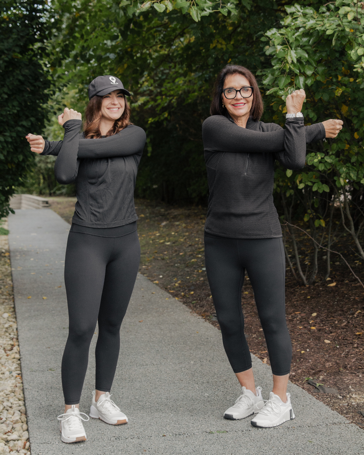 Caroline and Kelly from @Well stretching outdoors on a walking path surrounded by trees, demonstrating the importance of daily movement, mobility, and exercise for long-term health and longevity.