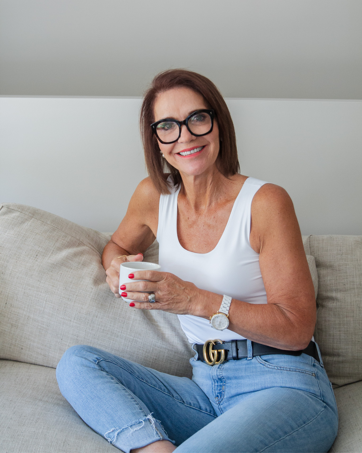 Kelly from @Well sitting on a neutral couch holding a coffee mug and smiling toward the camera in a bright, relaxed living room setting.