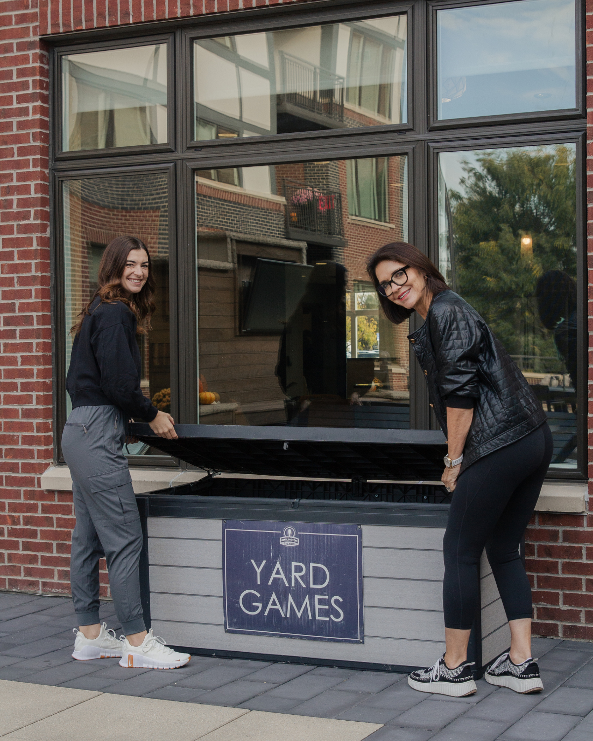 Caroline and Kelly open a box labeled yard games in the courtyard of their apartment complex.