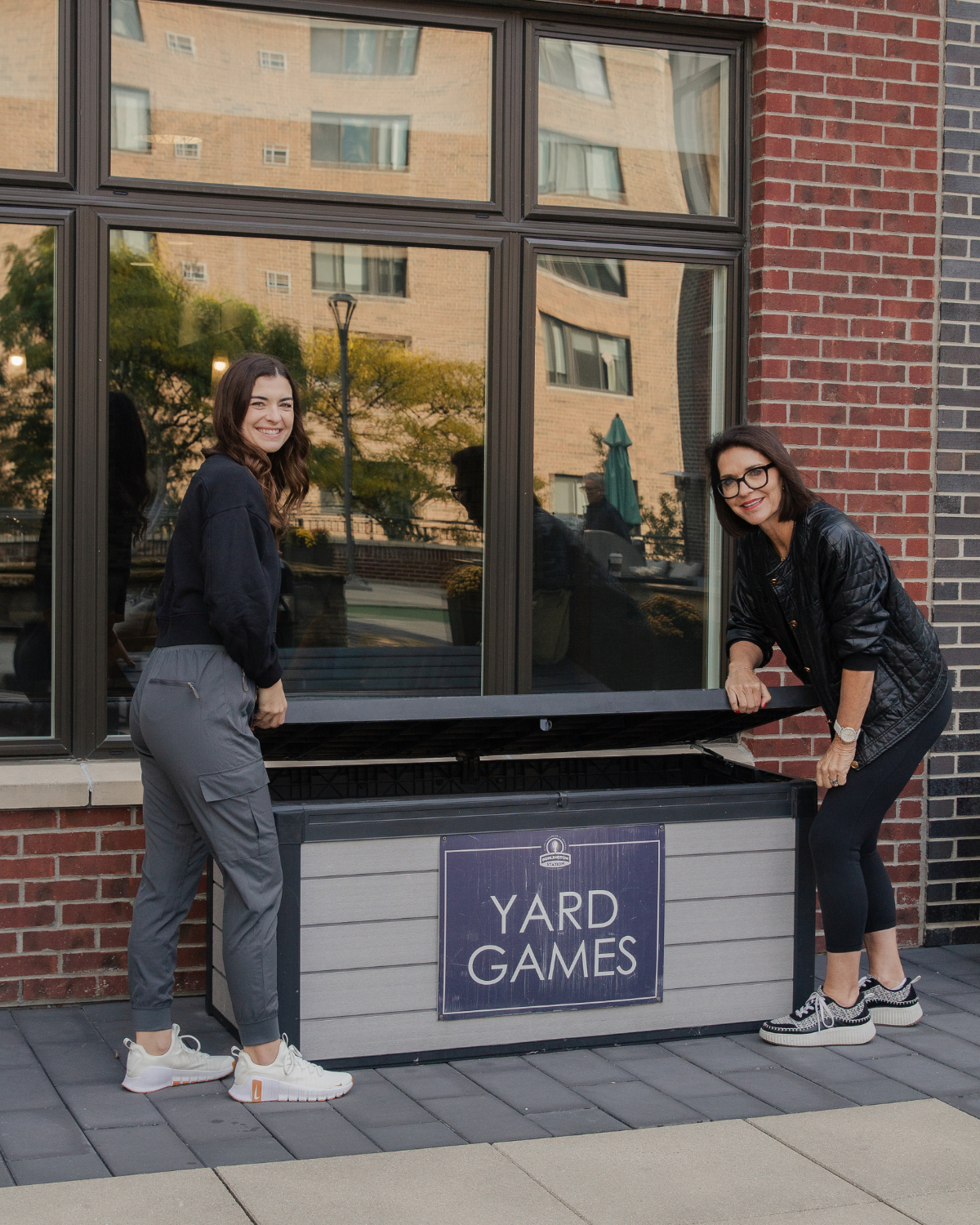 Kelly and Caroline standing beside a yard games storage box outdoors, highlighting fun and accessible ways to stay active through everyday movement and outdoor recreation.