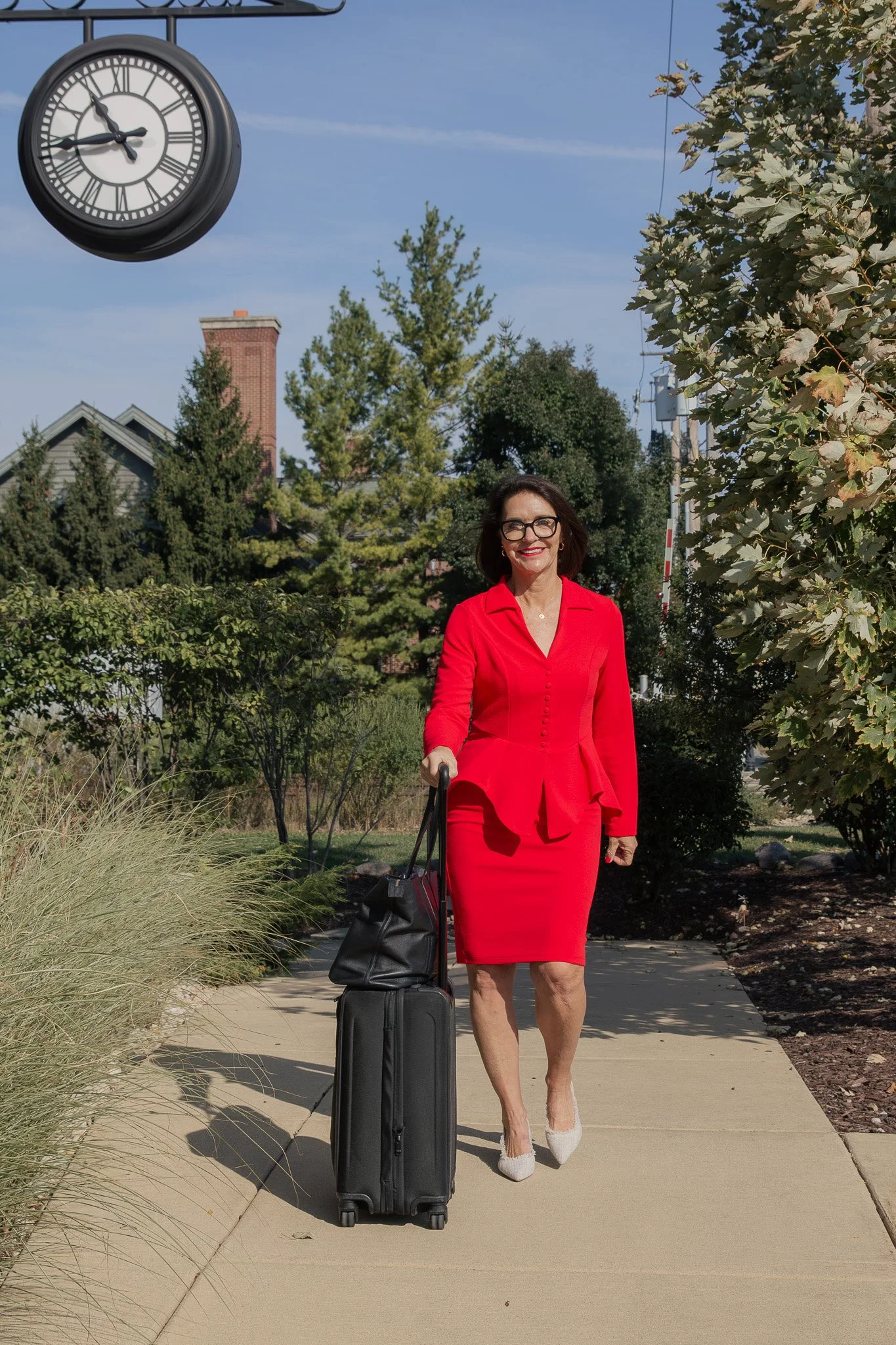 Kelly of @Well wearing a red business suit and walking outdoors with rolling luggage.