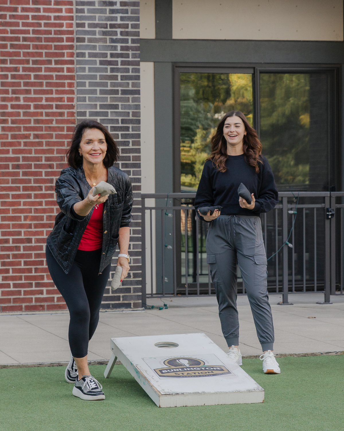 Two women playing cornhole outside during a community wellness gathering at Burlington Station, smiling and enjoying time together outdoors.