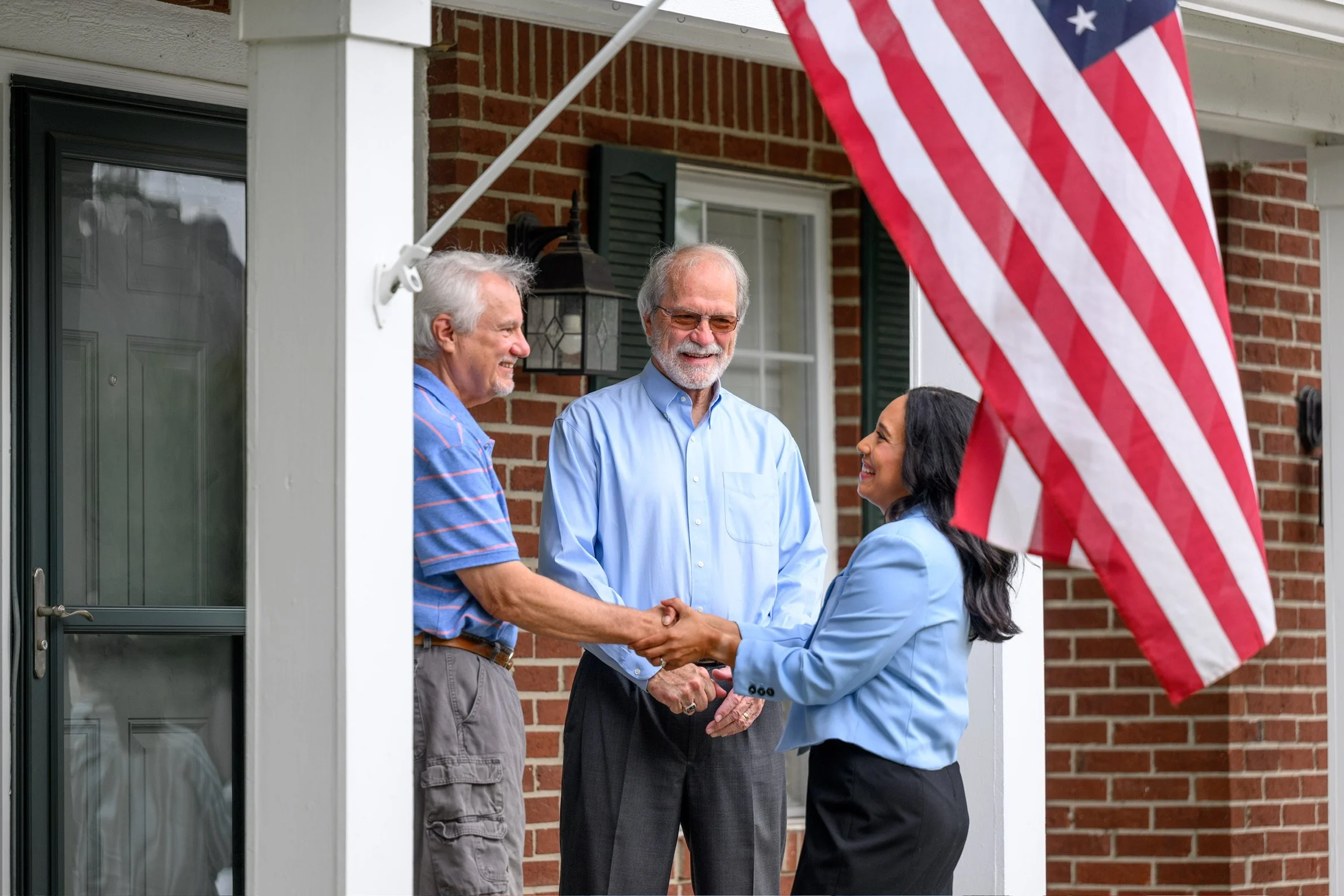 A woman in a blue blazer shaking hands with an older man on a porch, with two other men standing nearby, and an American flag in the foreground.