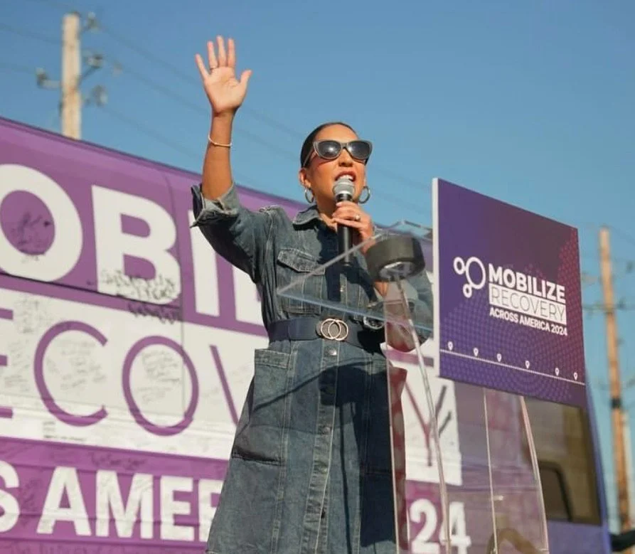 Woman speaking at a rally or event with a purple sign