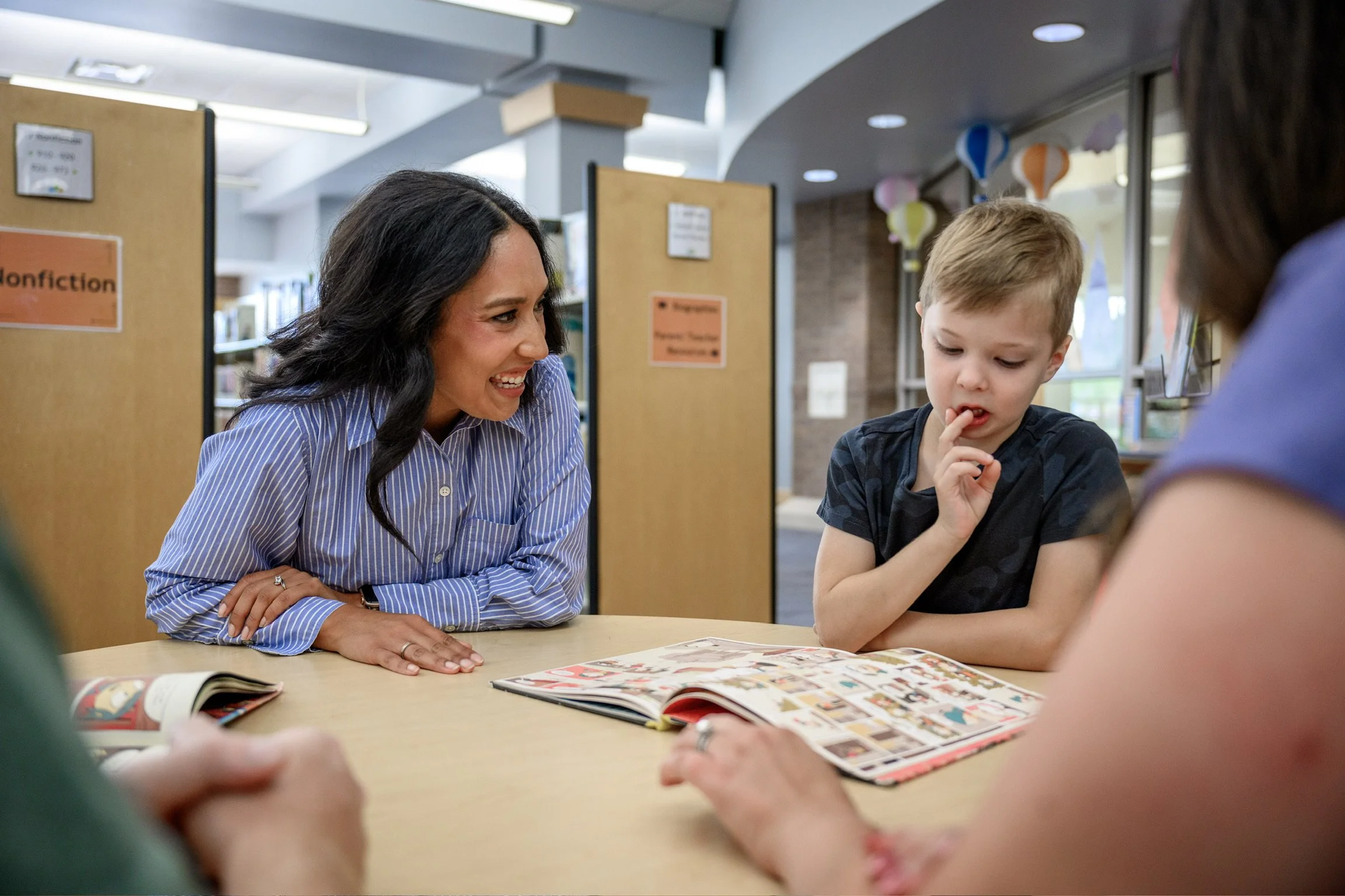 A woman with dark, wavy hair and a blue pinstripe shirt sitting at a table with a young boy with short, blond hair and a dark shirt, looking at comic books or magazines together in a library, with shelves and colorful lantern decorations in the background.