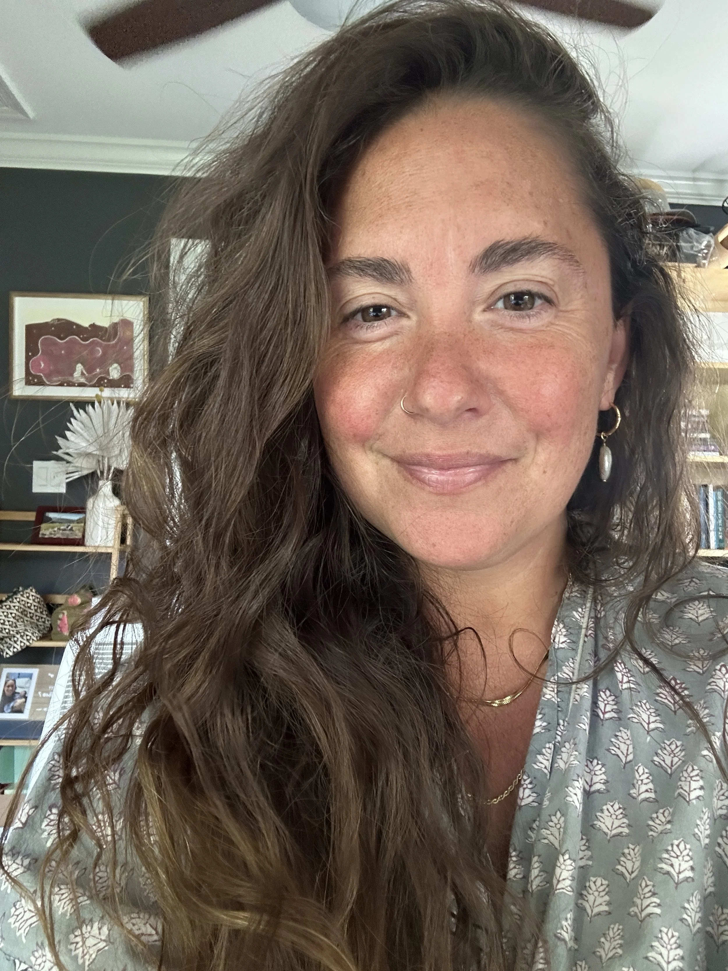 Smiling woman with long hair, wearing a printed blouse, gold earrings, and necklace, in a living room setting with art and books in the background.