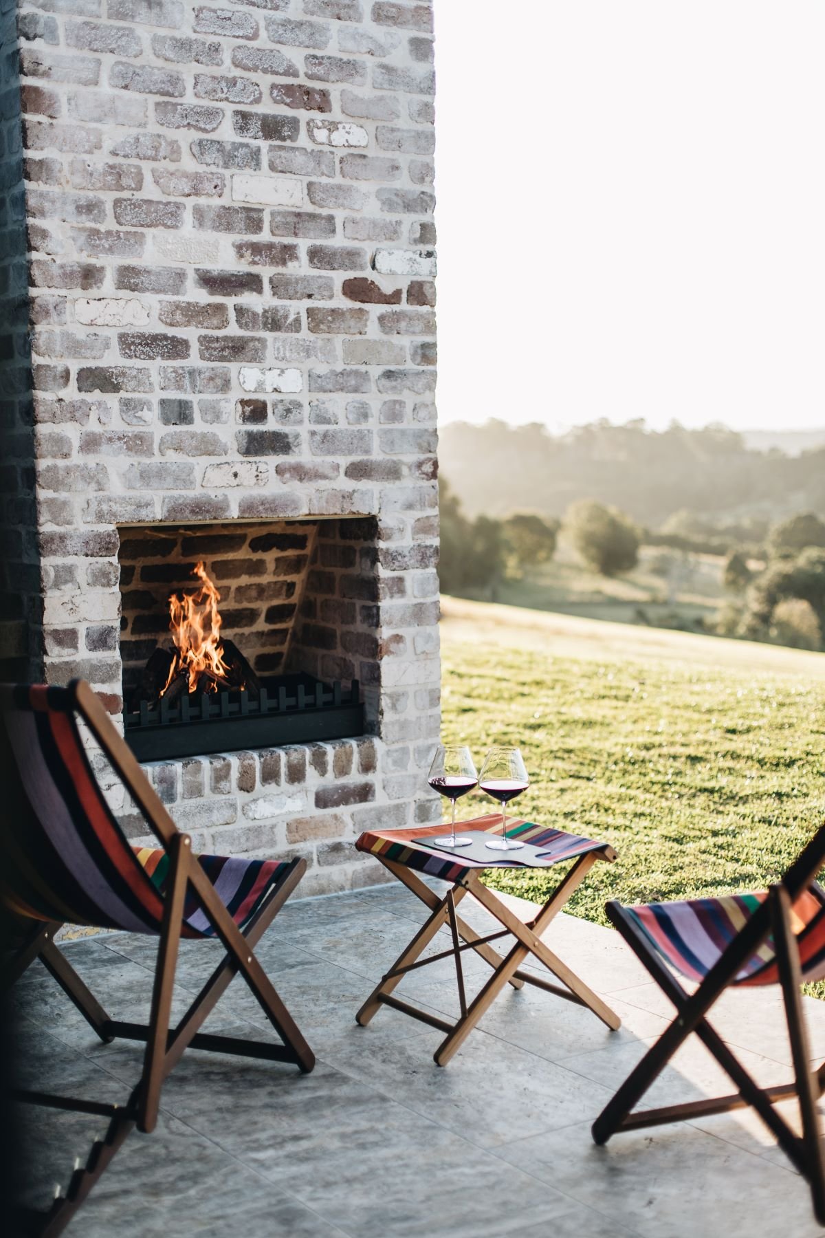 Outdoor patio with brick fireplace, two striped folding chairs, a small table with wine glasses, and a scenic countryside view.