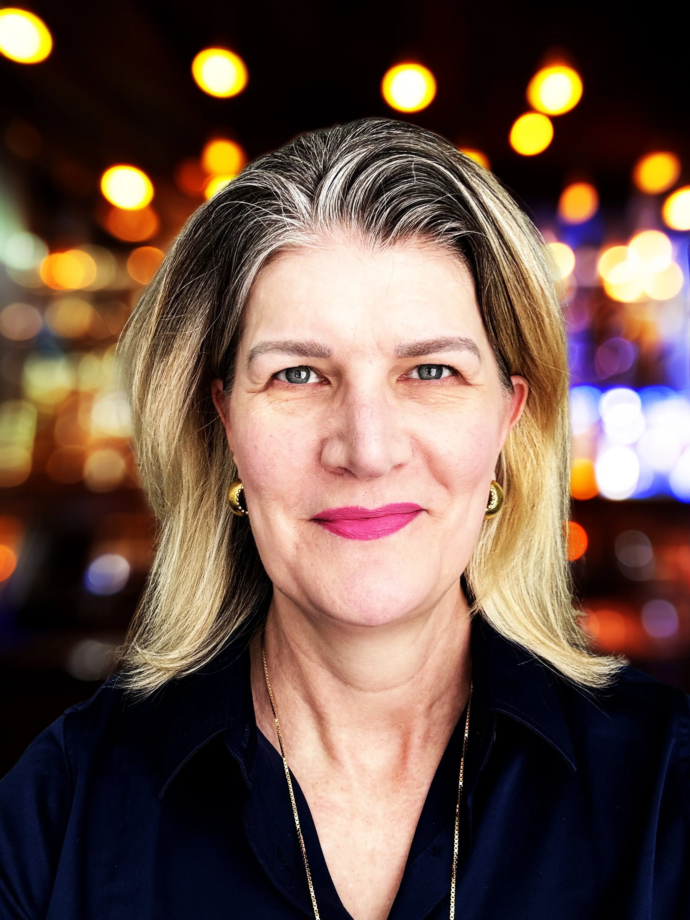 A woman with shoulder-length blonde hair, wearing pink lipstick, gold earrings, and a navy blouse, smiling with a blurred colorful bokeh background.