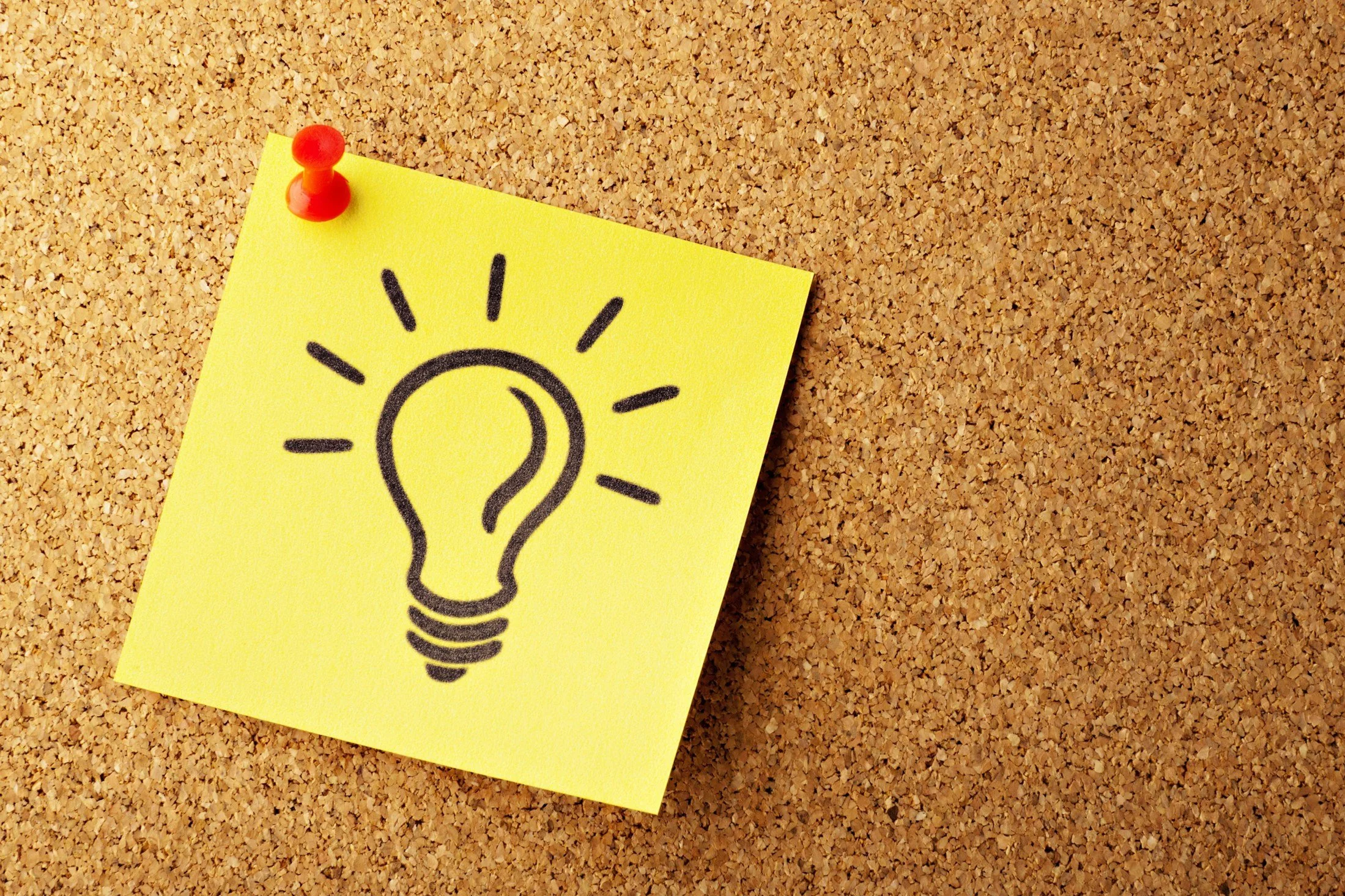 Yellow sticky note with a black drawing of a light bulb, pinned to a cork bulletin board - get your business copywriting and content sorted.