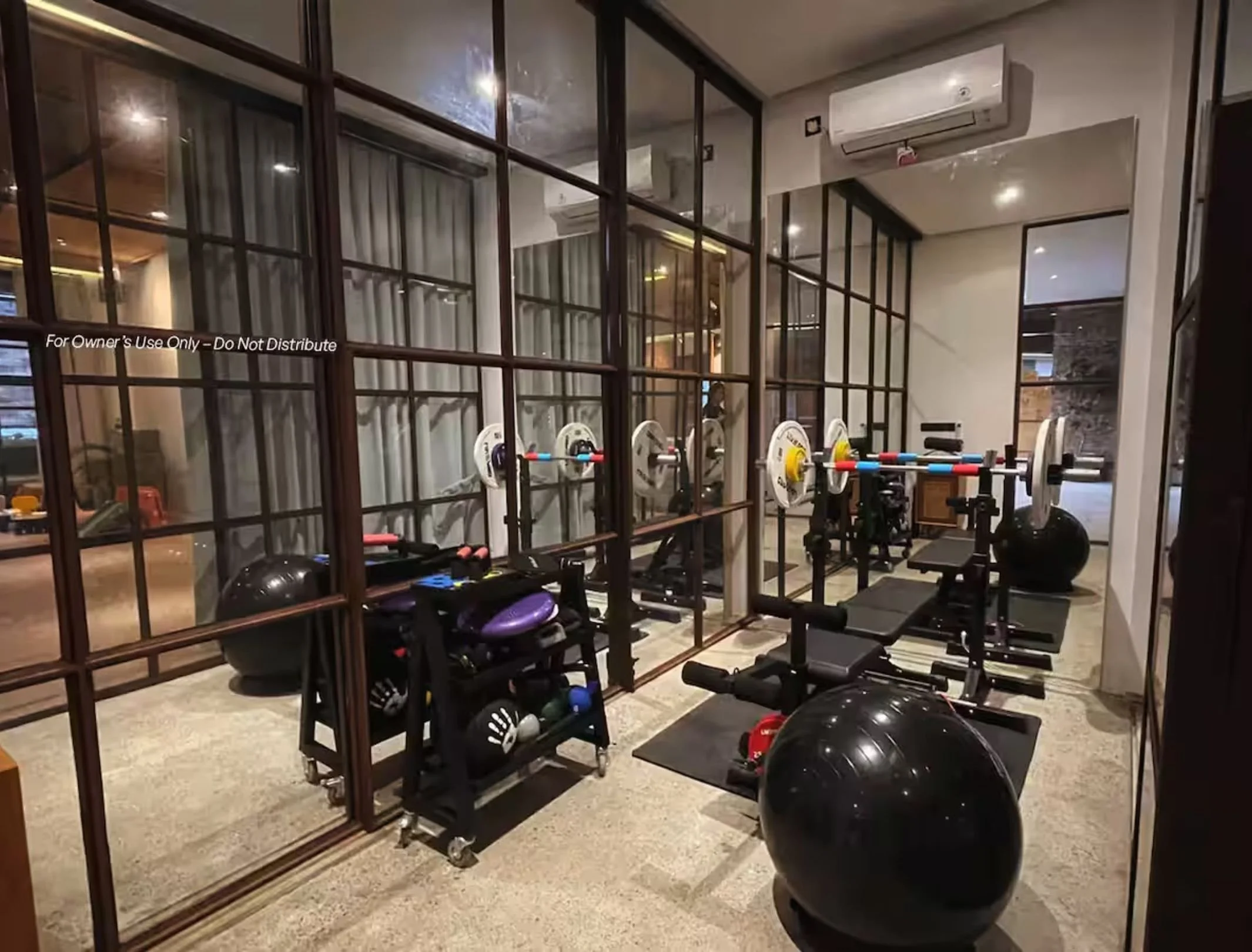 Gym workout area with weightlifting equipment, including a barbell with weights on a bench, large exercise ball, dumbbells, and a weight rack, separated by glass partitions in an indoor fitness space.