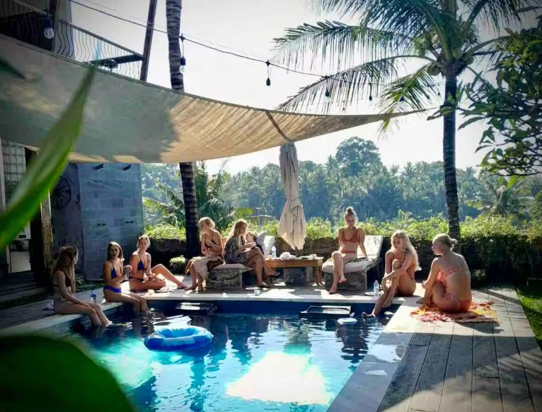 People in swimsuits relaxing by a small outdoor pool surrounded by tropical trees and plants, with lounge chairs and shaded area.