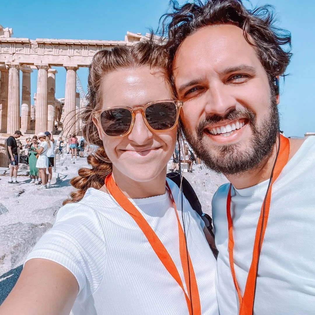 A smiling couple taking a selfie in front of the Parthenon ruins in Athens, Greece, on a sunny day. The woman is wearing sunglasses and a white shirt, and the man has a beard and curly hair, both with orange lanyards around their necks.