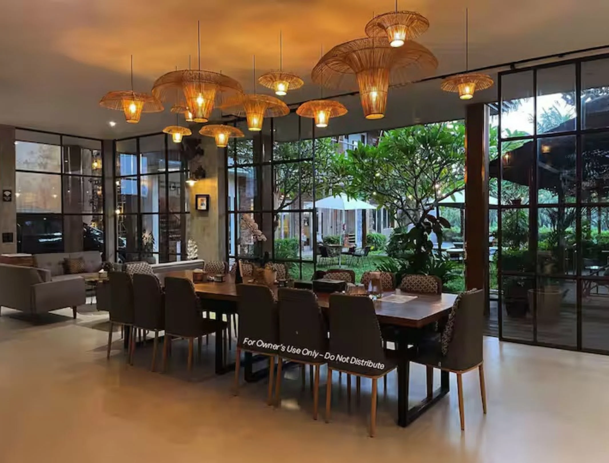 Elegant dining area with large wooden table, surrounded by upholstered chairs, inside a restaurant with floor-to-ceiling windows revealing a lush outdoor garden, with bamboo-style pendant lights hanging from the ceiling.