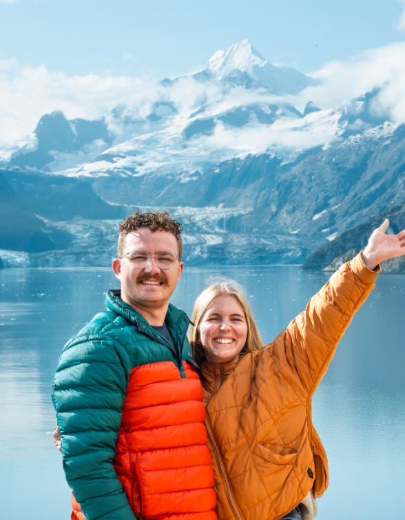 A smiling man and woman in colorful winter jackets standing by a lake with snow-capped mountains in the background. The woman is raising her arm in excitement.