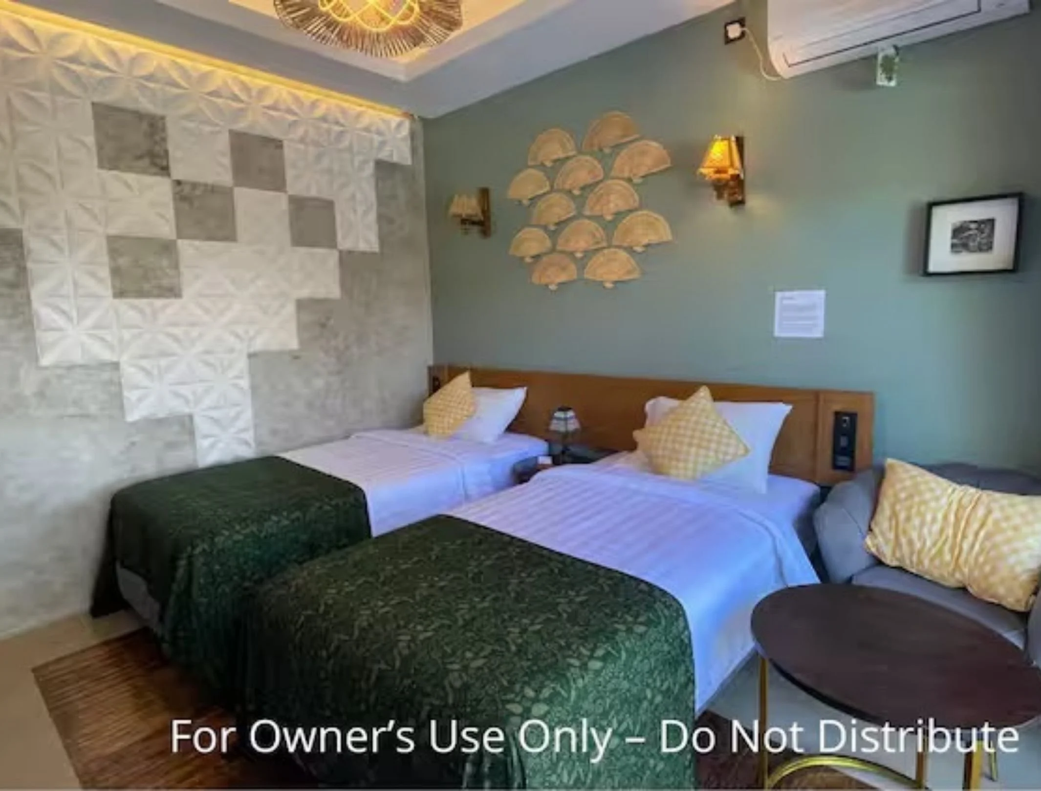 A hotel room with two twin beds, decorated with yellow checkered pillows and green bedspreads. There is a small sitting area with a round table and a gray couch with a yellow checkered pillow. The wall behind the beds has decorative wall art and wall-mounted lamps, and the room features part of an air conditioning unit.