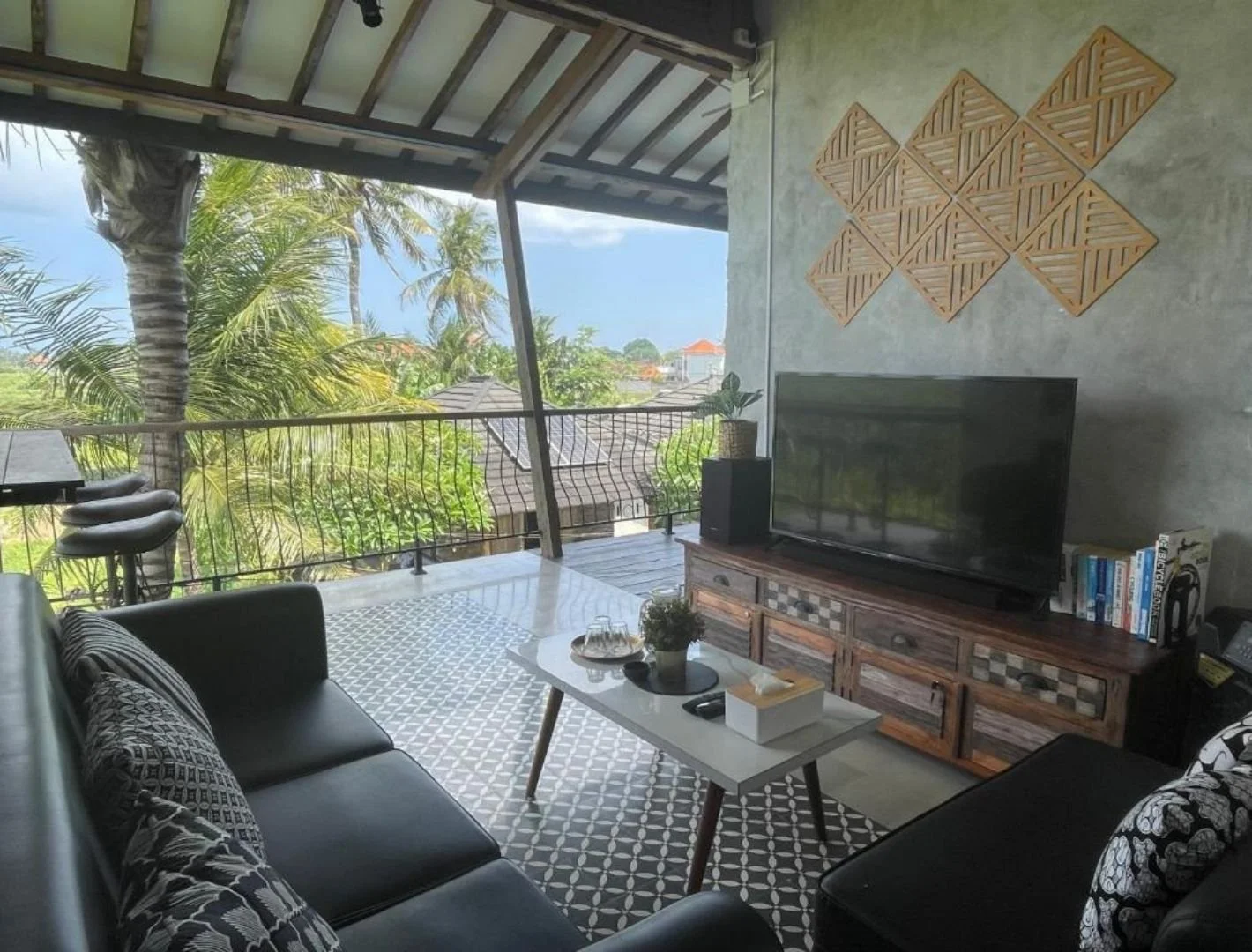 Living room with open balcony view of tropical trees, sofa with patterned cushions, coffee table with plant and tissue box, wooden TV stand with TV, bookshelf, and wall decor