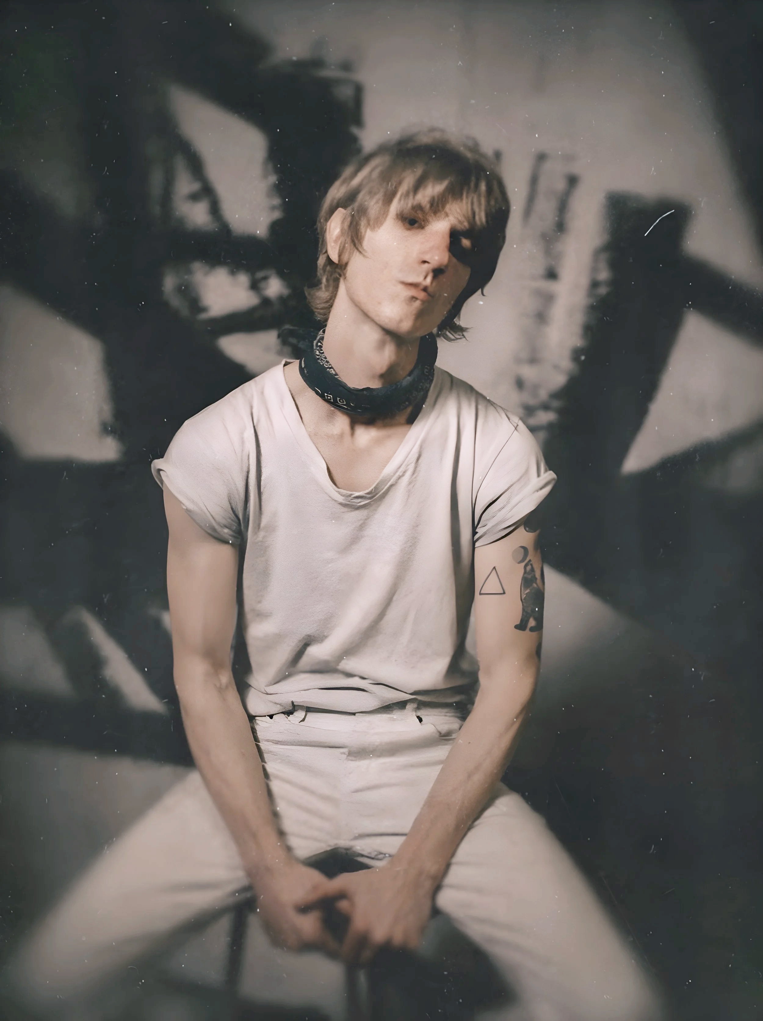 A young man with light brown hair, wearing a white t-shirt and white pants, sitting in front of a dark, abstract background with shadowy shapes. He has a bandana around his neck, a tattoo of a bear on his left arm, and is looking slightly away from the camera.