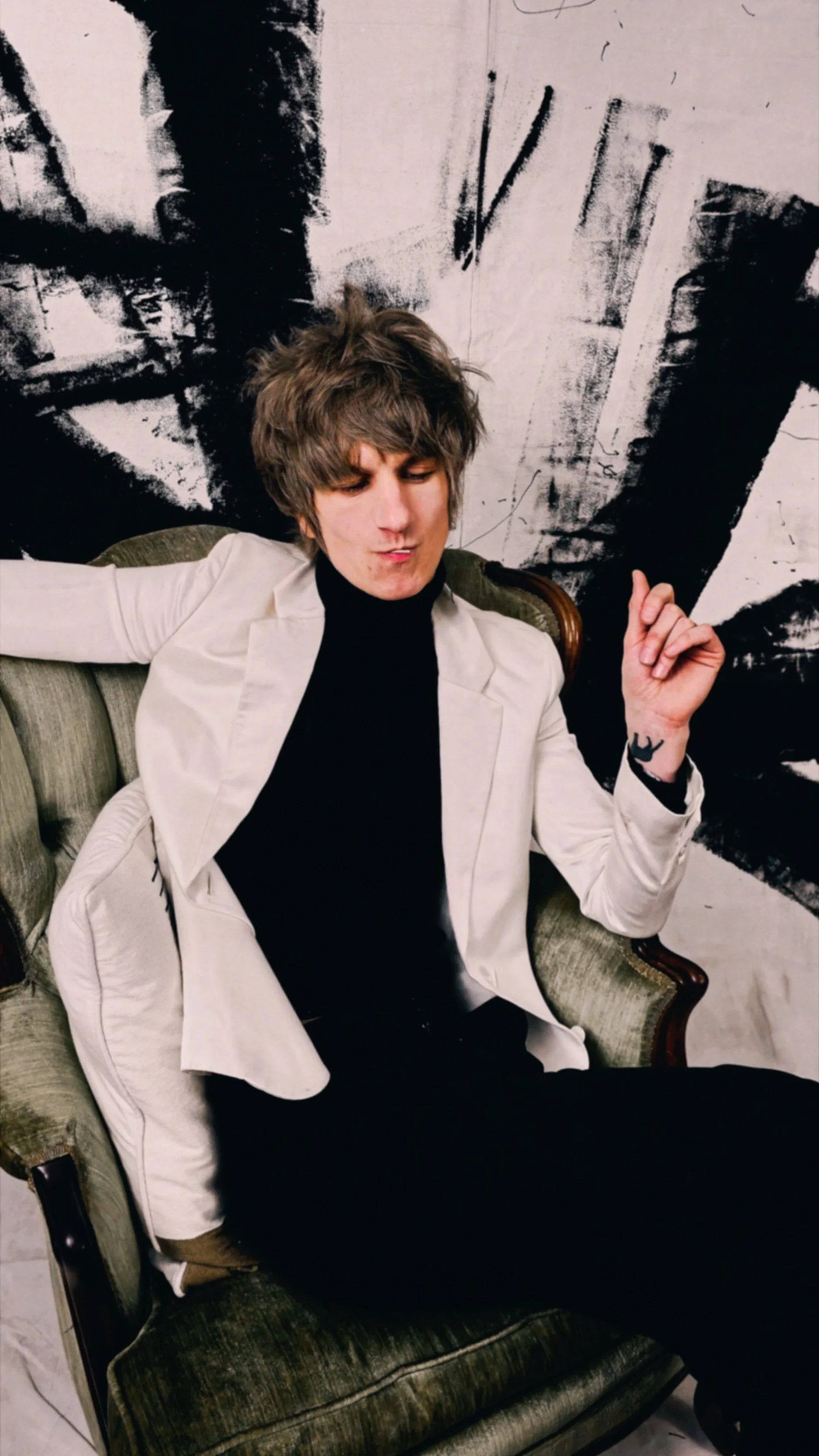A young man with messy hair, wearing a white blazer over a black turtleneck, is sitting on an antique green velvet armchair. He is displaying a relaxed, confident pose with one arm stretched out and a hand raised, with closed eyes and a slight smile, in front of a black-and-white abstract mural.