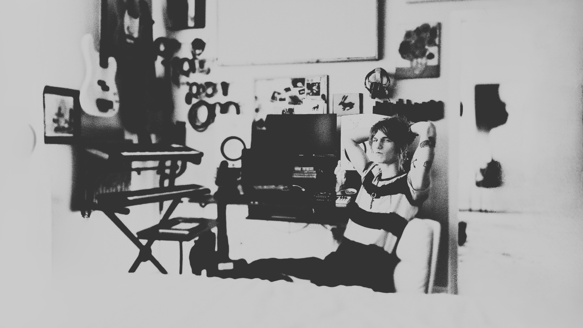 Black and white photo of a man with short hair and tattoos, sitting in a room with various objects, including a computer, paintings, and musical instruments, in the background.
