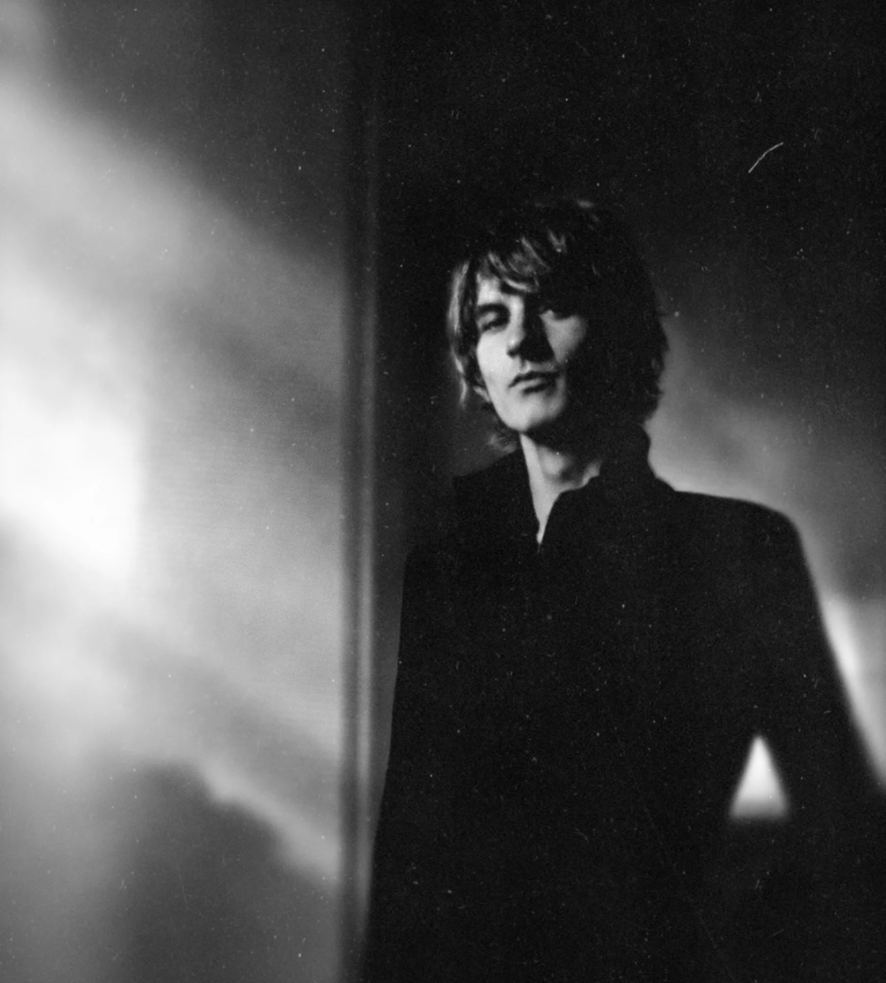 Black and white photo of a person with curly hair, with closed eyes, in front of a dark background. The person appears to be in a contemplative or relaxed state.