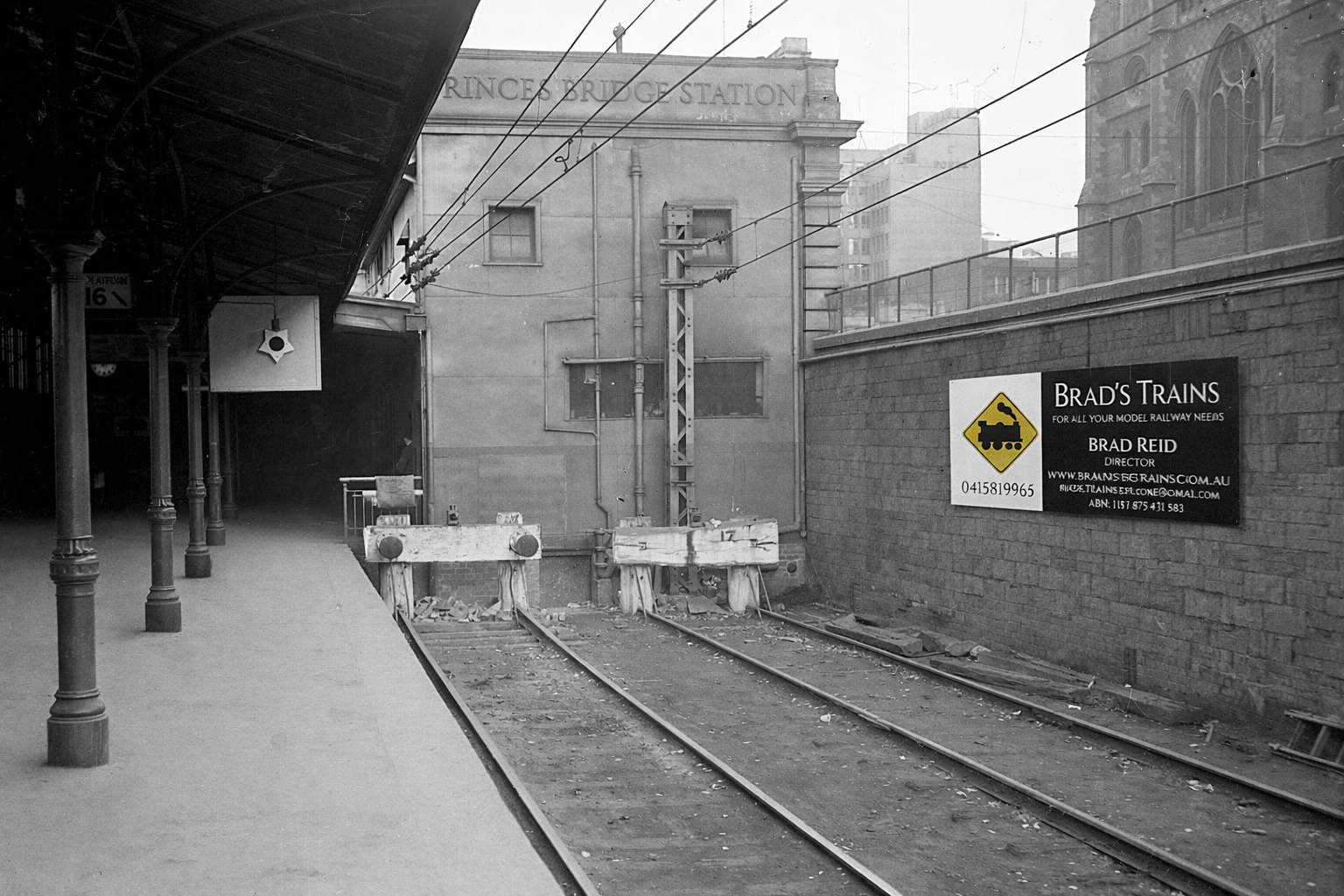 A deserted train station platform with a set of railway tracks ahead. There is a construction barrier on the tracks. The platform has a covered waiting area with columns. A sign on the wall reads 'Princes Bridge Station'. Another sign advertises 'Brad's Trains' with contact information, and includes a yellow warning triangle with a train symbol.