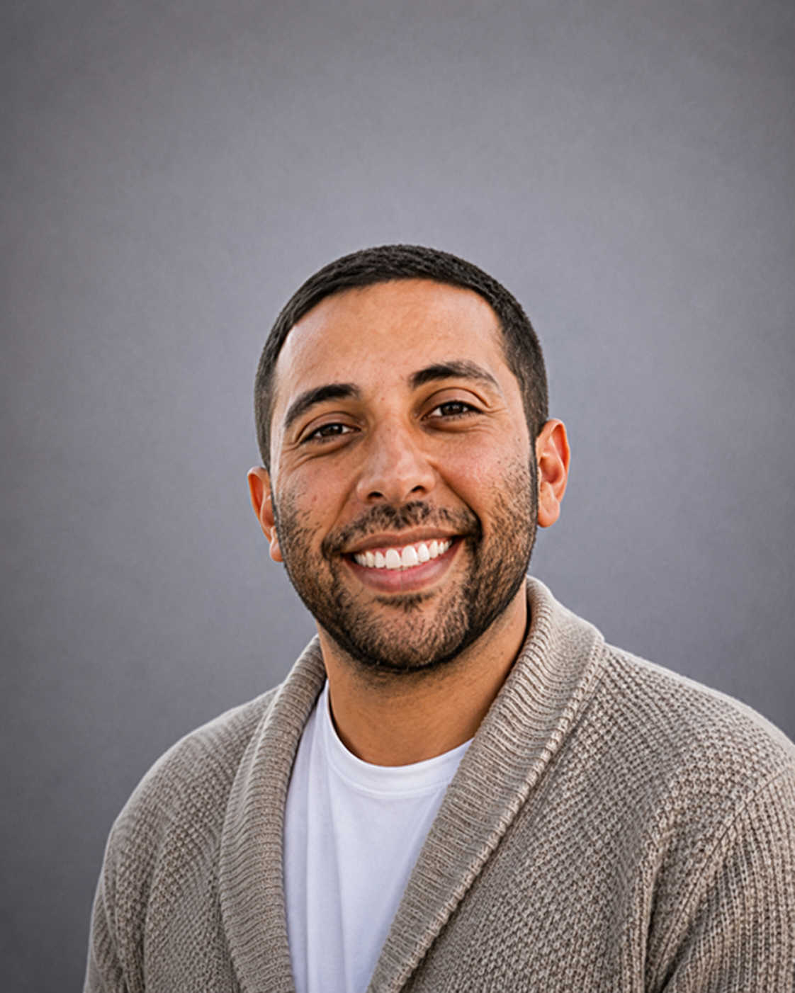 Portrait of a smiling man with short dark hair and beard wearing a beige cardigan and white t-shirt against gray background.