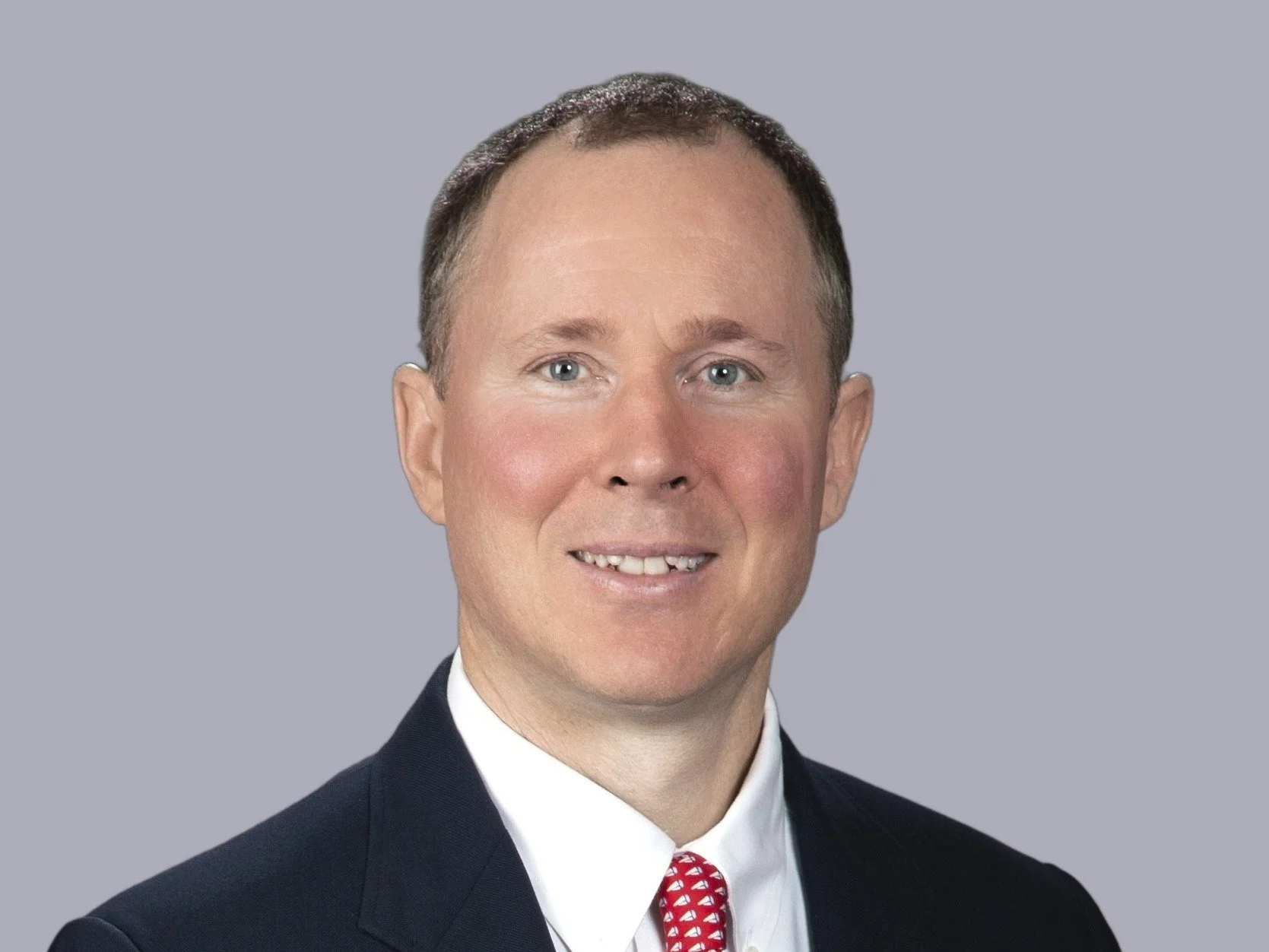 Headshot of a smiling man with short hair wearing a navy suit, white shirt, and red patterned tie against a grey background.