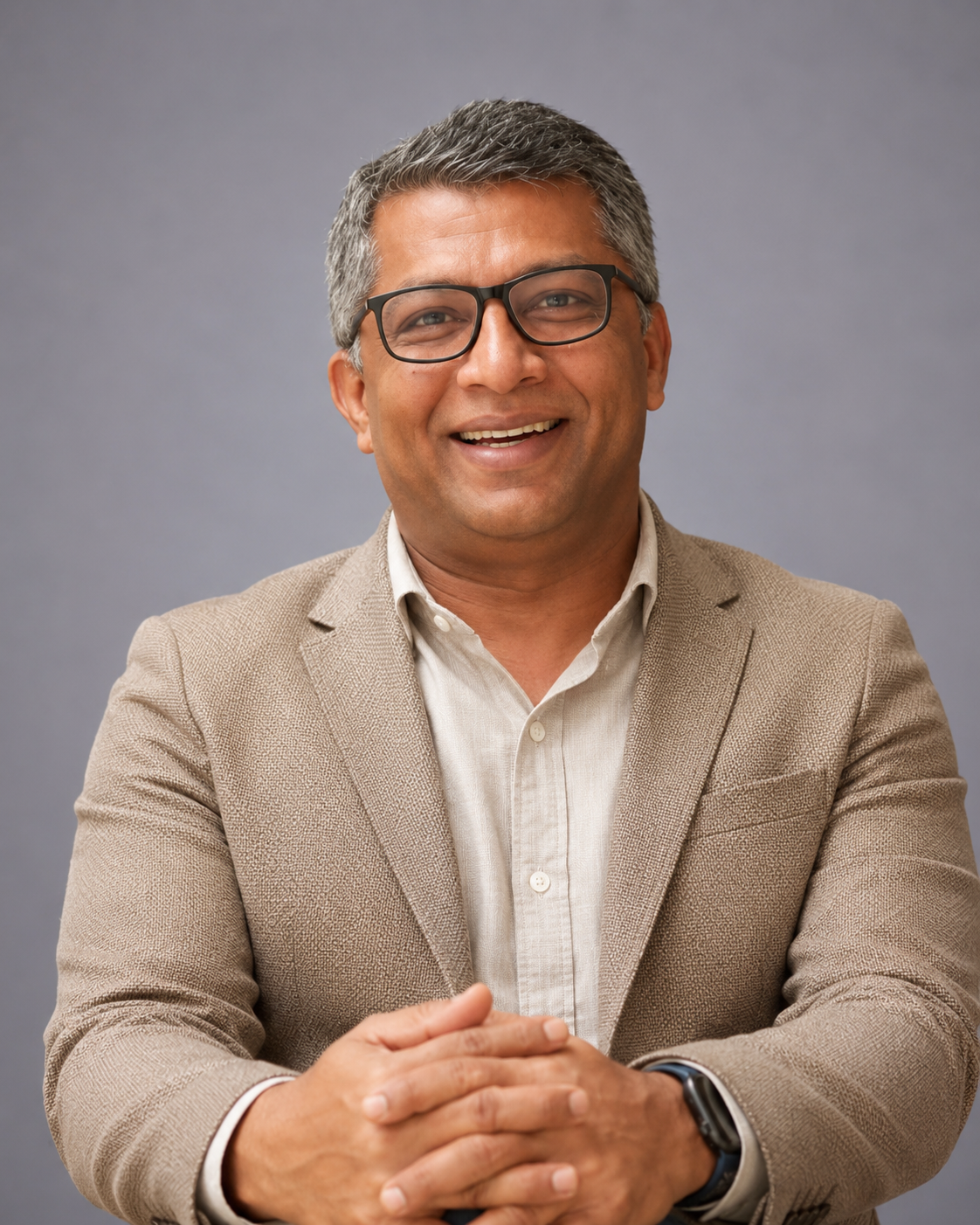 A man with short gray hair, wearing glasses, a beige blazer, and a white shirt, smiling with arms crossed, against a gray background.