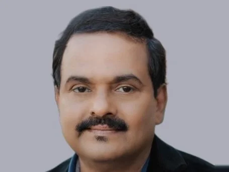 Headshot of a man with dark hair and a mustache wearing a dark jacket over a blue collared shirt against a grey background.
