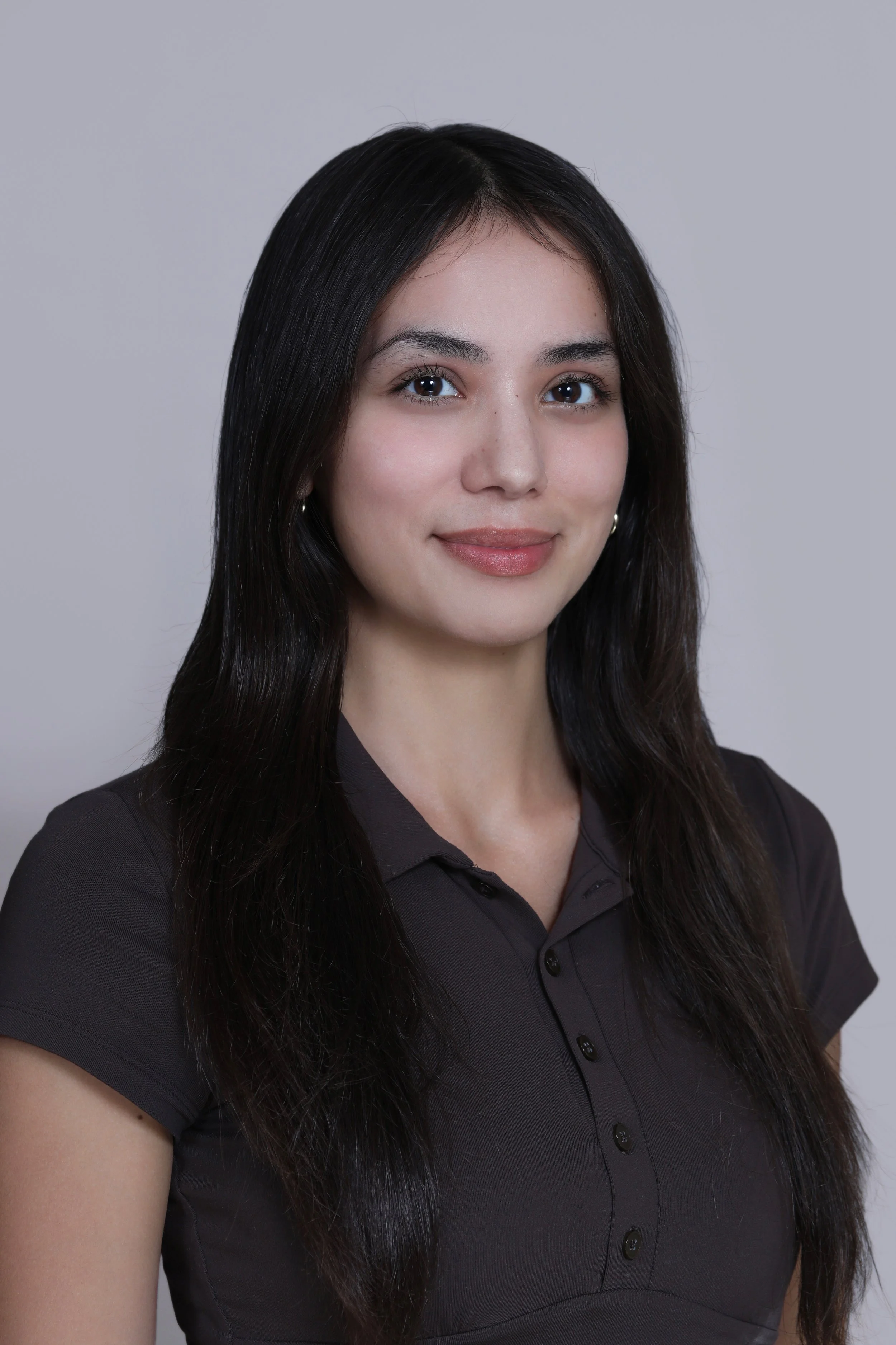 Portrait of a young woman with long dark hair, smiling, wearing a dark button-up shirt, against a plain gray background.