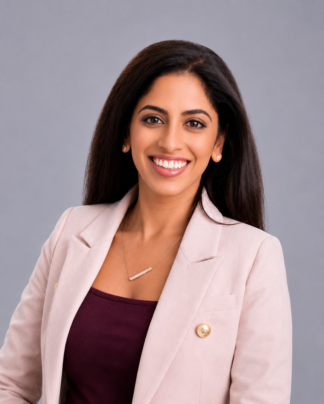 A woman with long dark hair, wearing a beige blazer with a gold pin, a burgundy top, and a necklace, smiling against a gray background.