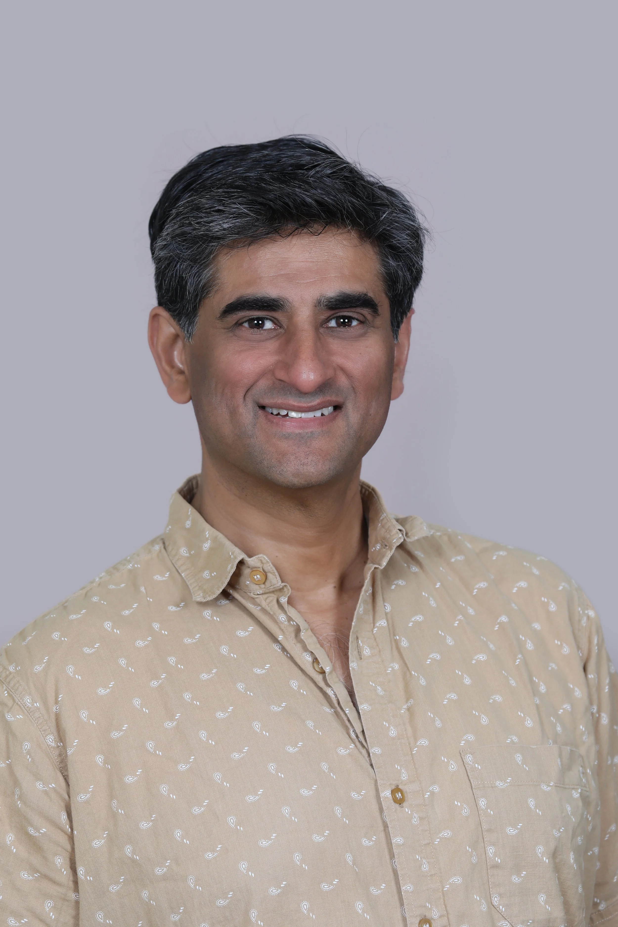 A man with dark hair and a light skin tone is smiling and looking at the camera. He is wearing a beige button-up shirt with a white pattern, set against a plain gray background.